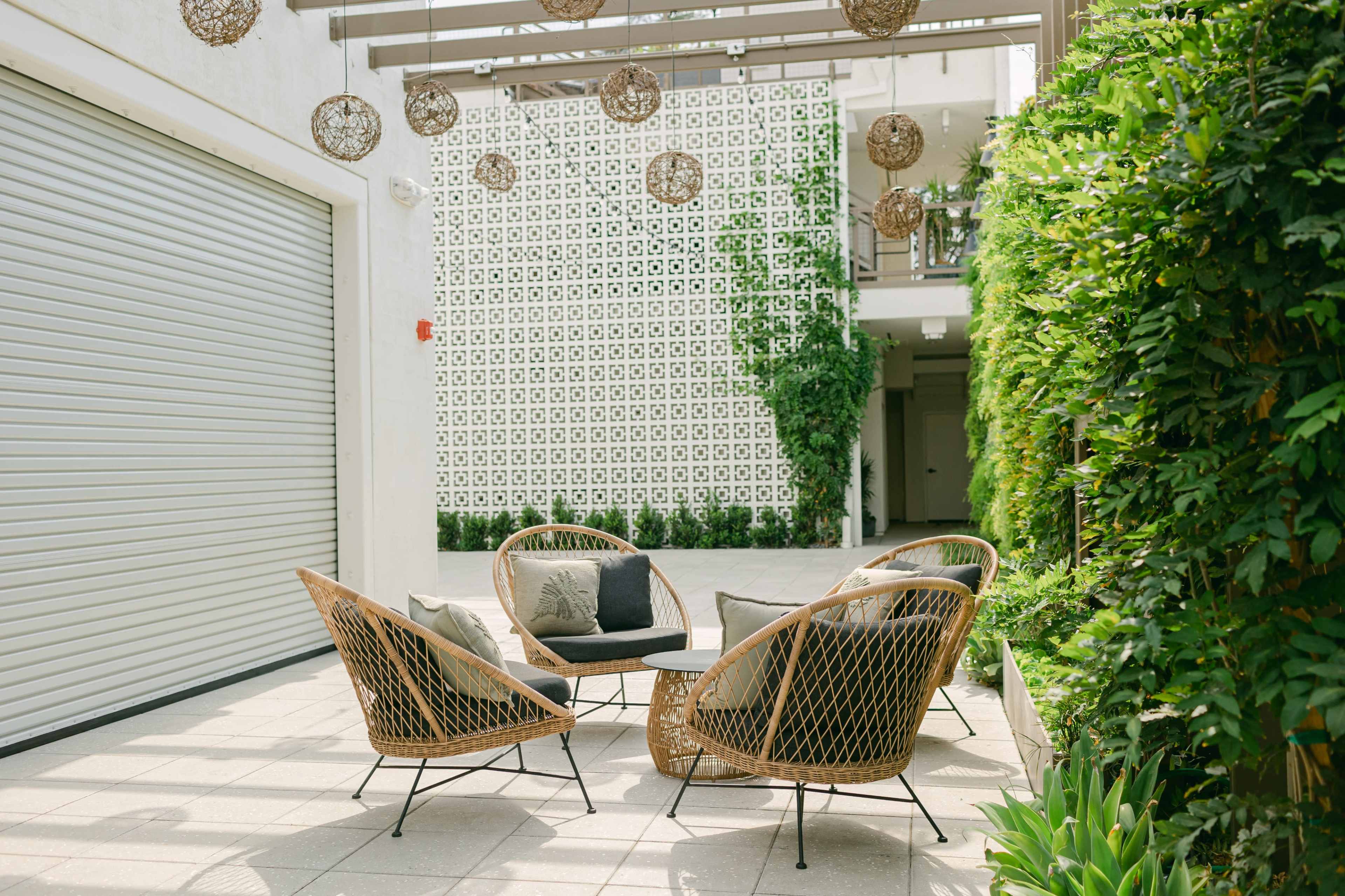 A modern outdoor seating area features four woven chairs surrounding a small table, with greenery and a patterned wall in the background.