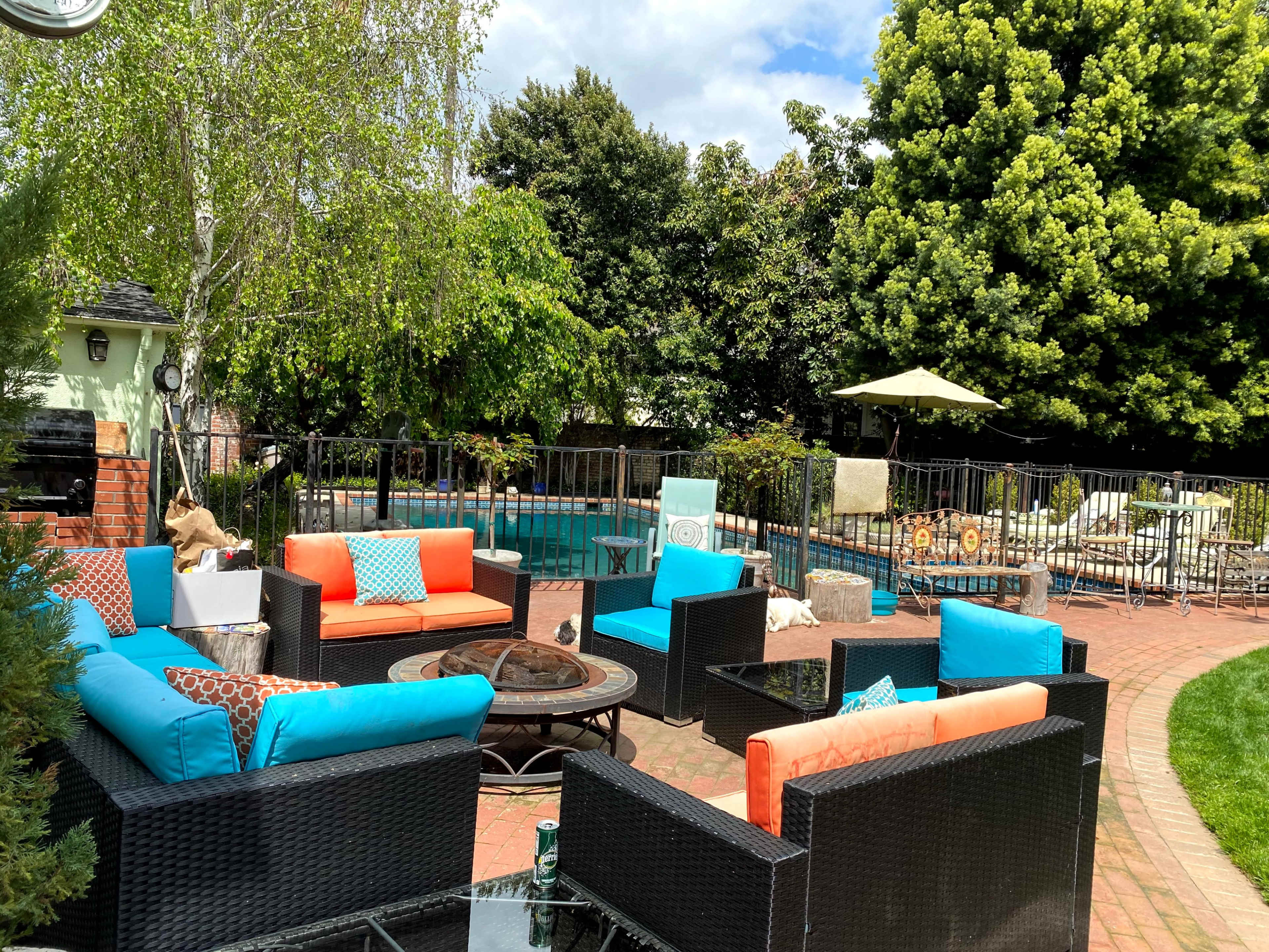 A landscaped backyard with a swimming pool surrounded by seating areas featuring bright blue and orange cushions, along with a fire pit and lush green trees.