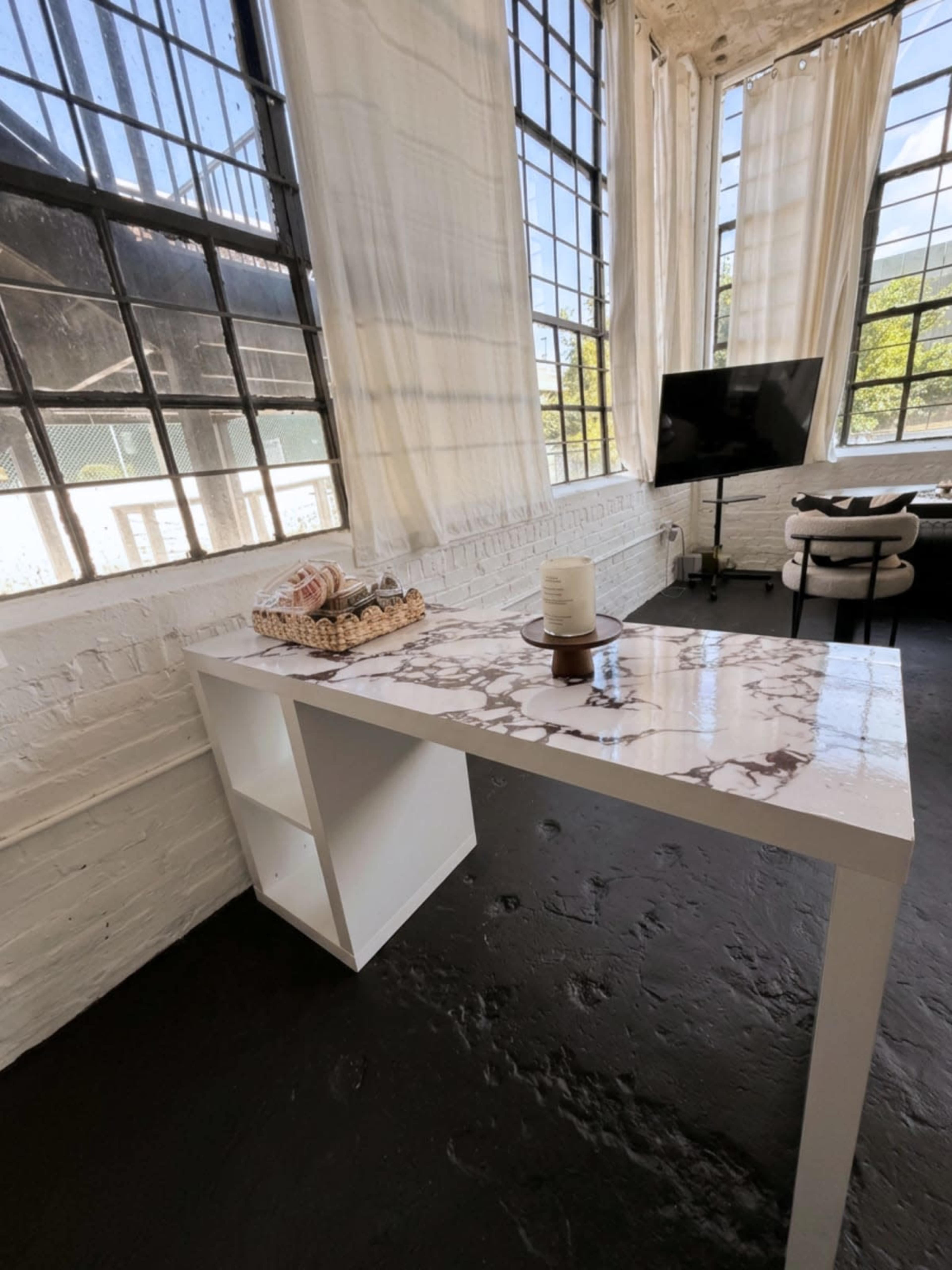 The image shows a modern white desk with a marble-patterned top, positioned near large industrial-style windows in a bright interior space.