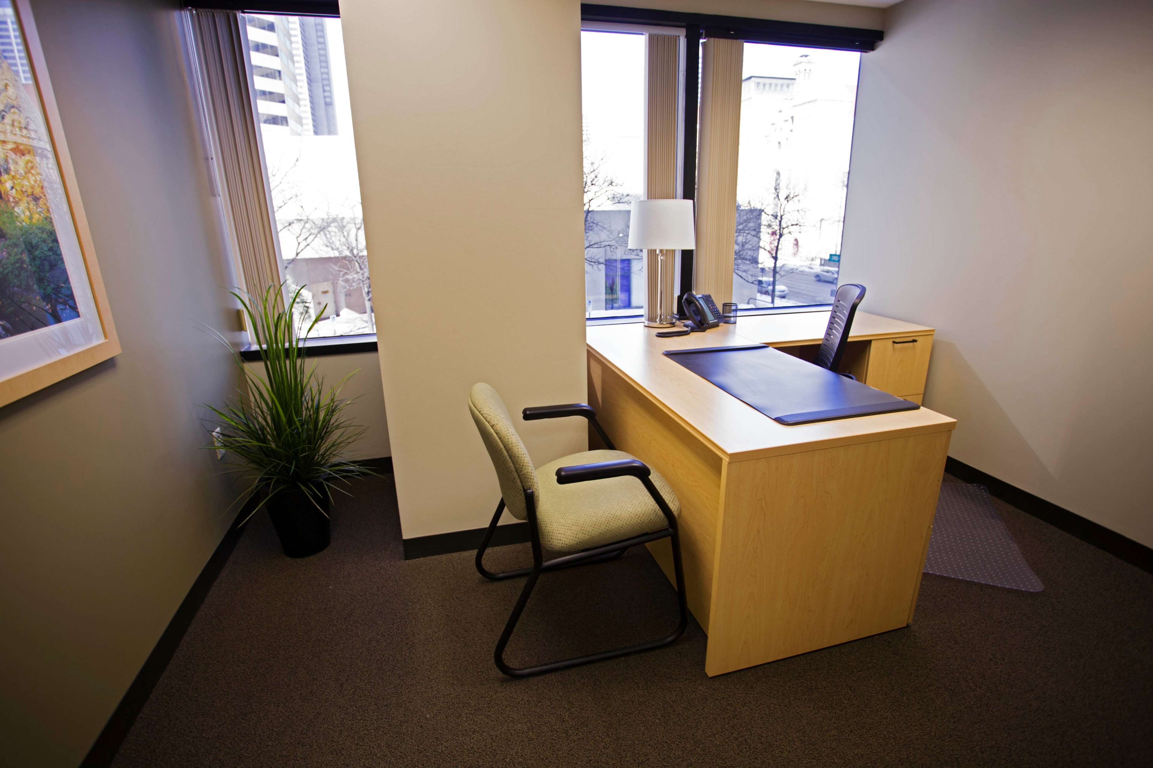 An office space features a wooden desk with a phone, a lamp, and two chairs near large windows.