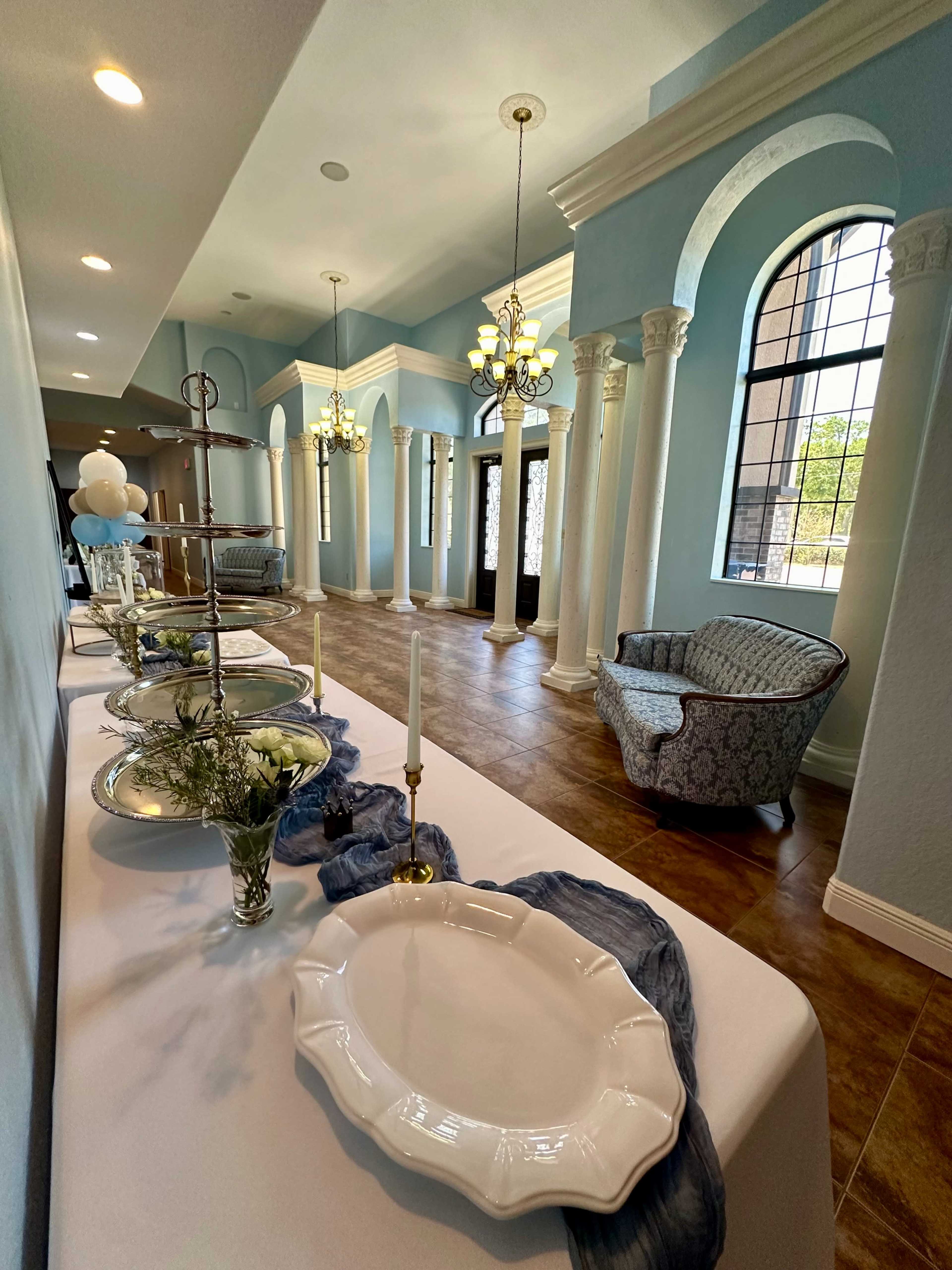 The image shows a decorated hallway with elegant columns, a table set with dishes and candles, and a seating area with a patterned chair.