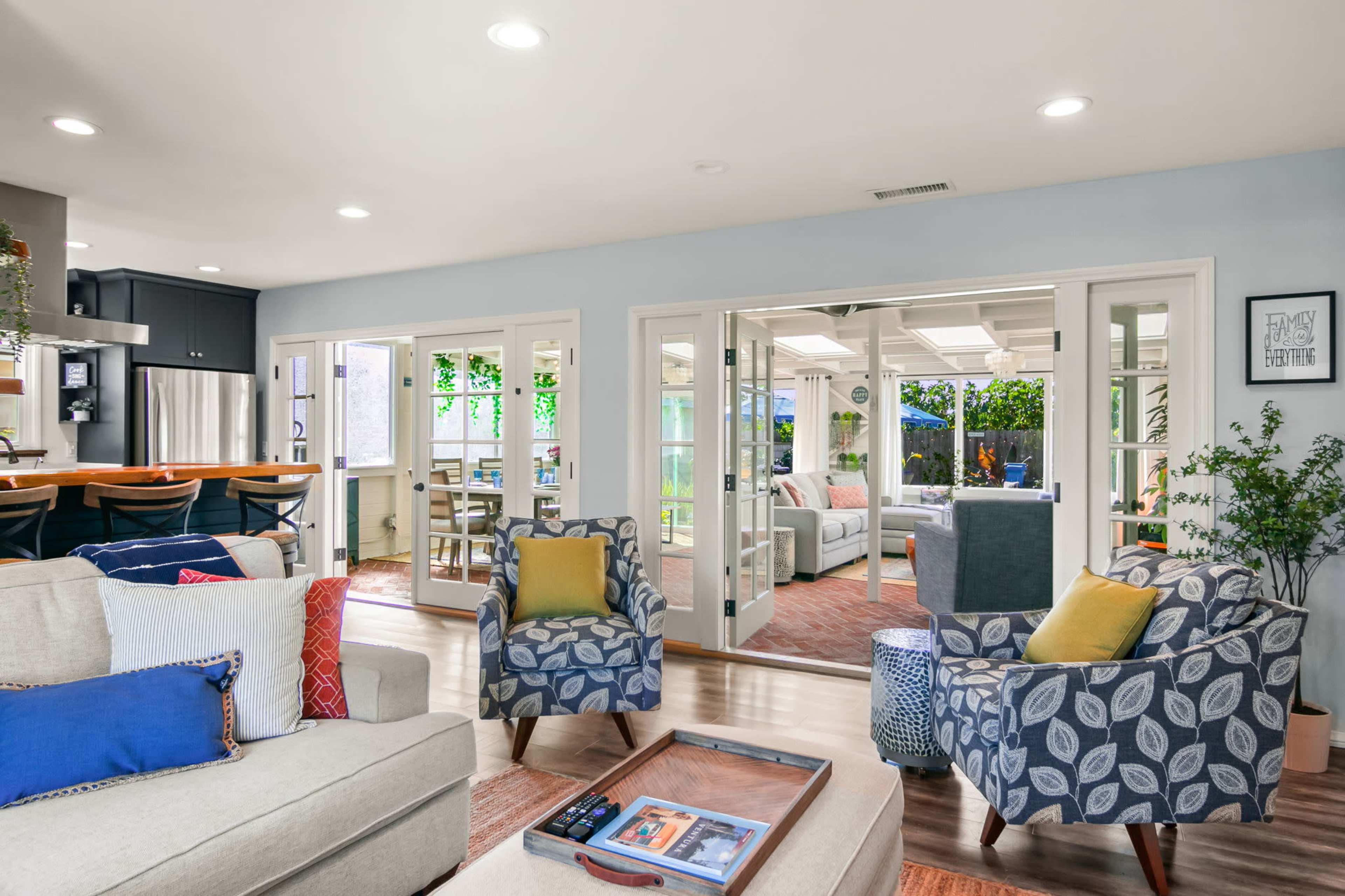 The image shows a modern open-concept living room with comfortable seating, a wooden coffee table, and large glass doors leading to a sunlit patio area.