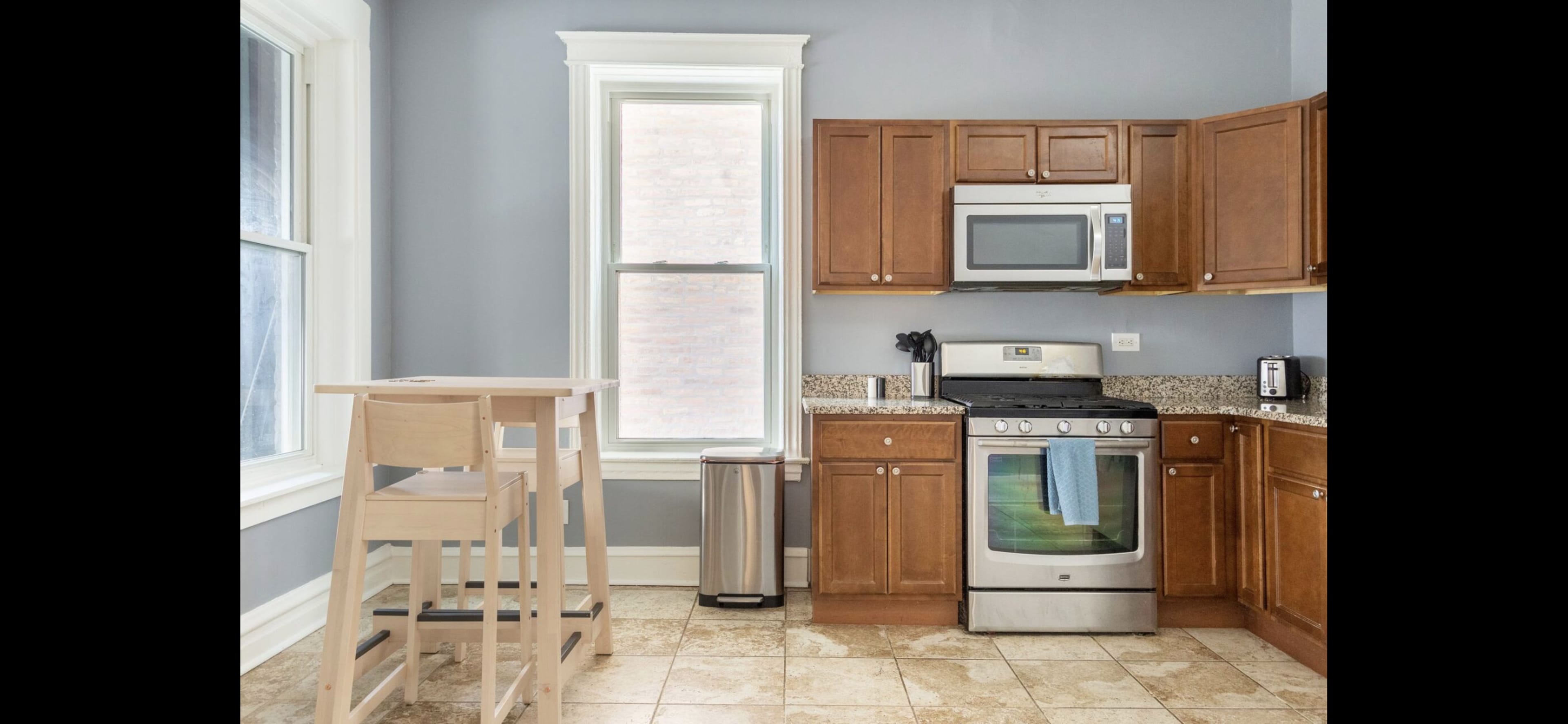 A kitchen features wooden cabinets, a stainless steel stove, a microwave, and a small wooden table with two chairs beside a window.