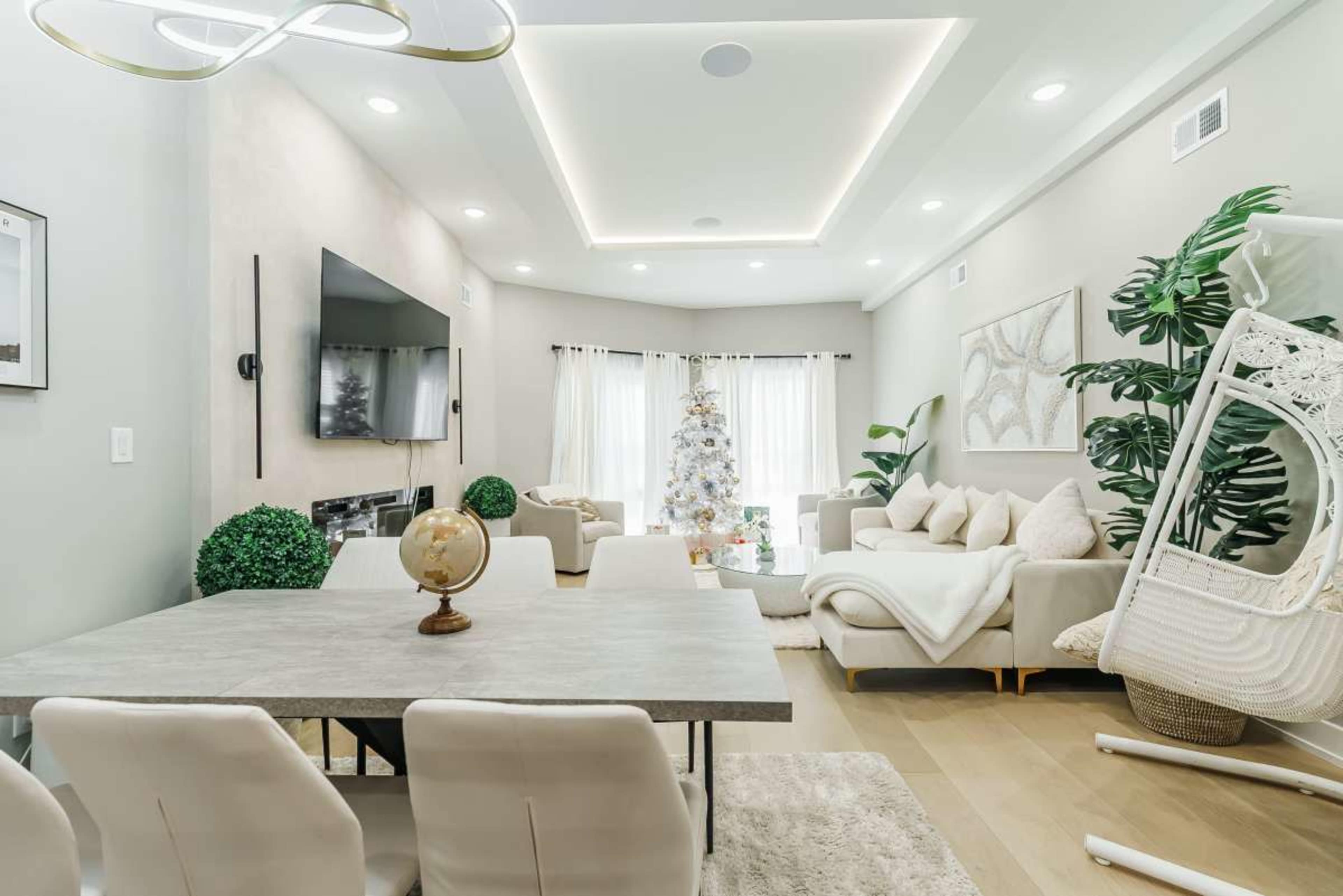 The image shows a modern living room and dining area decorated with a Christmas tree, surrounded by light-colored furniture and indoor plants.