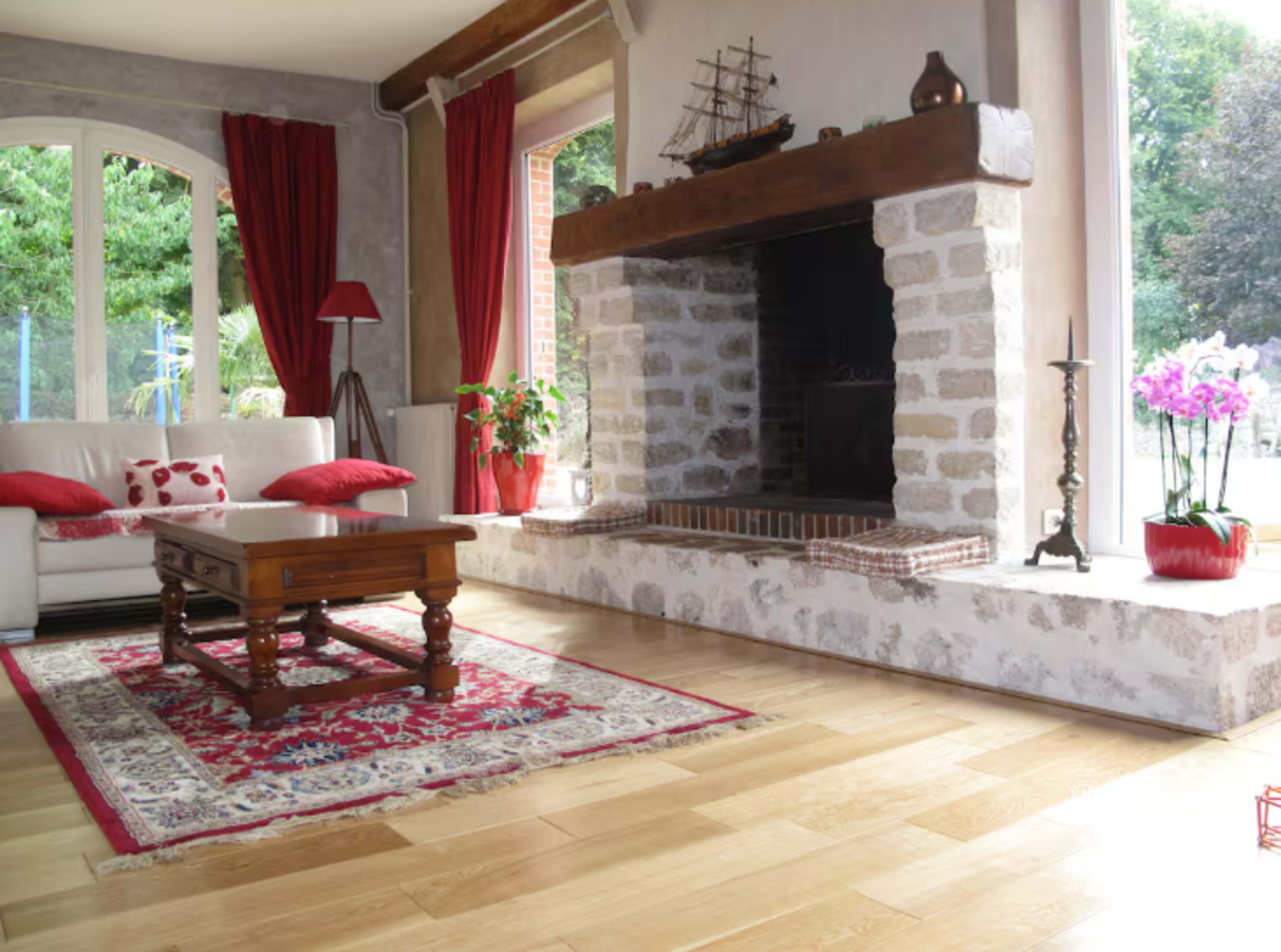 The image shows a sunlit living room with a stone fireplace, wooden coffee table, and a white couch adorned with red cushions.
