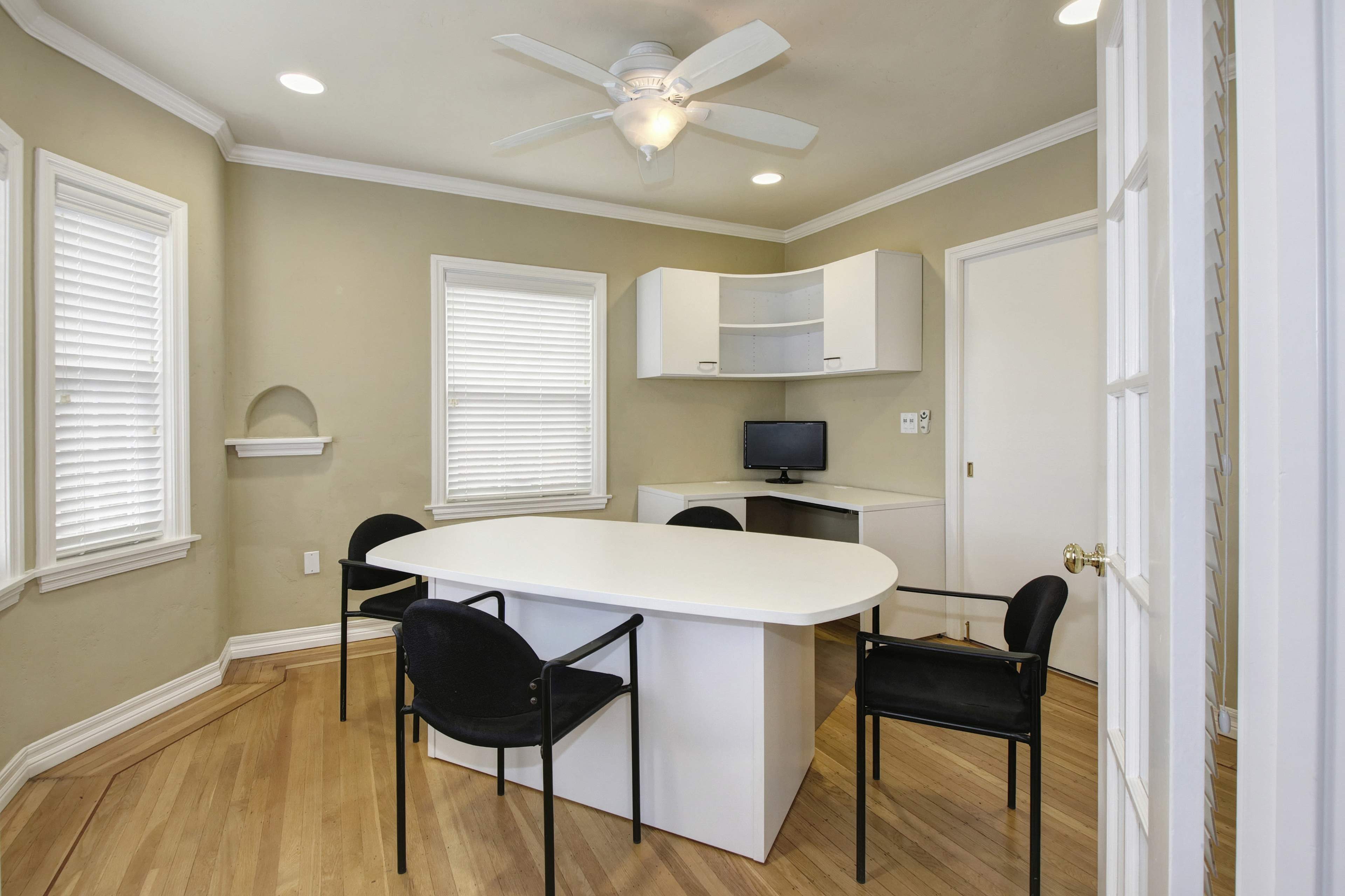 The image depicts a small workspace with a round table, four black chairs, a wall-mounted desk with a computer, and natural light from multiple windows.