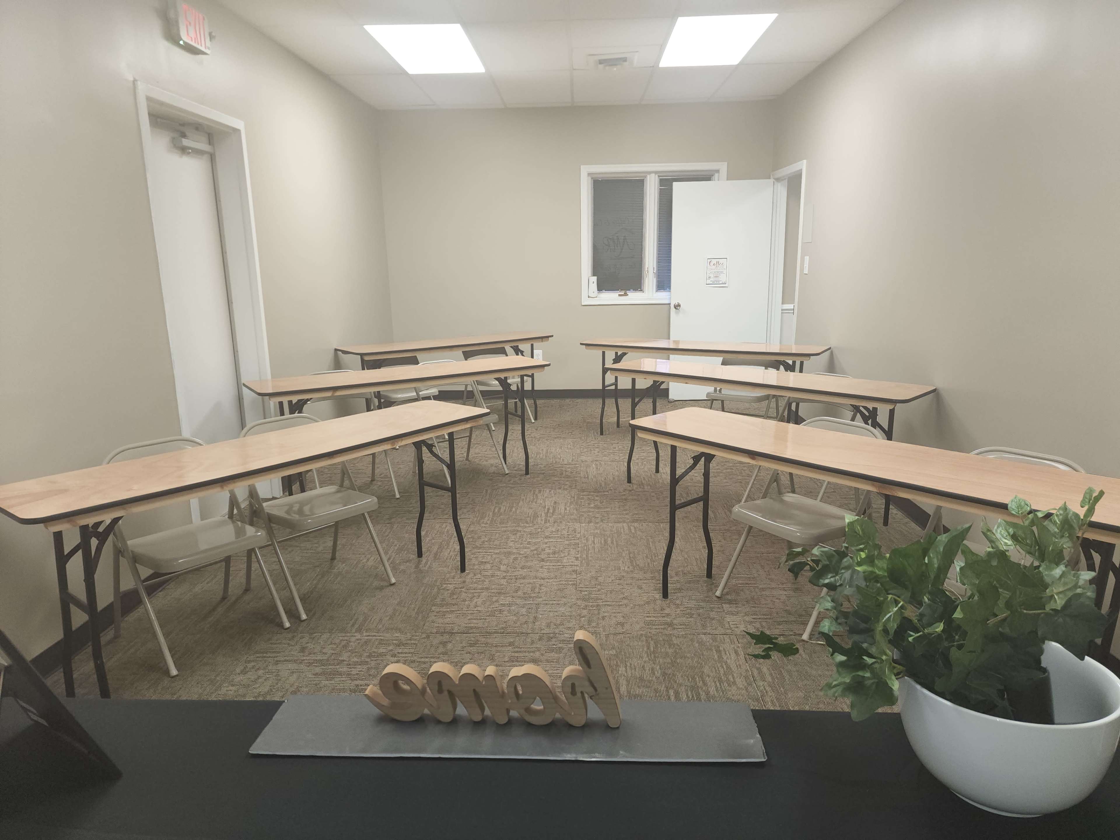 A small, vacant classroom with several tables arranged in a U-shape and a plant on a desk at the front.