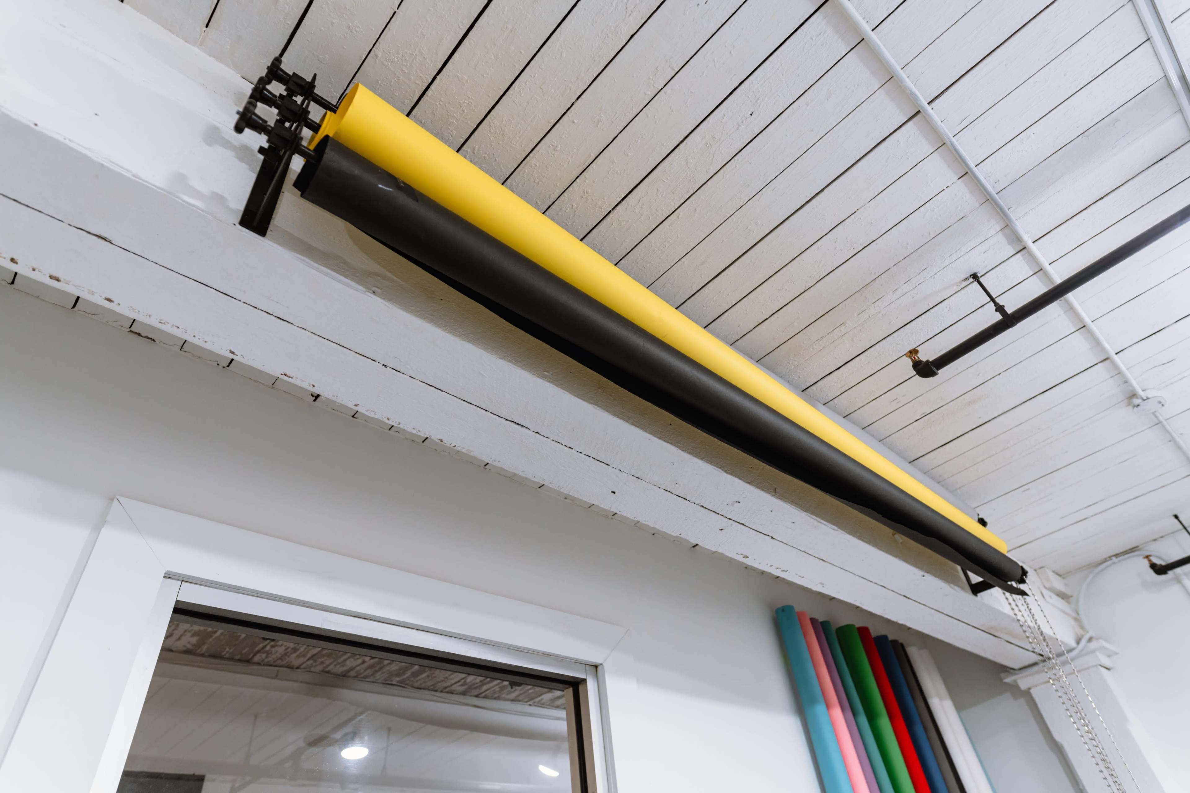The image shows yellow and black paper rolls mounted on a ceiling above a window, with various colorful rolls visible in the background.