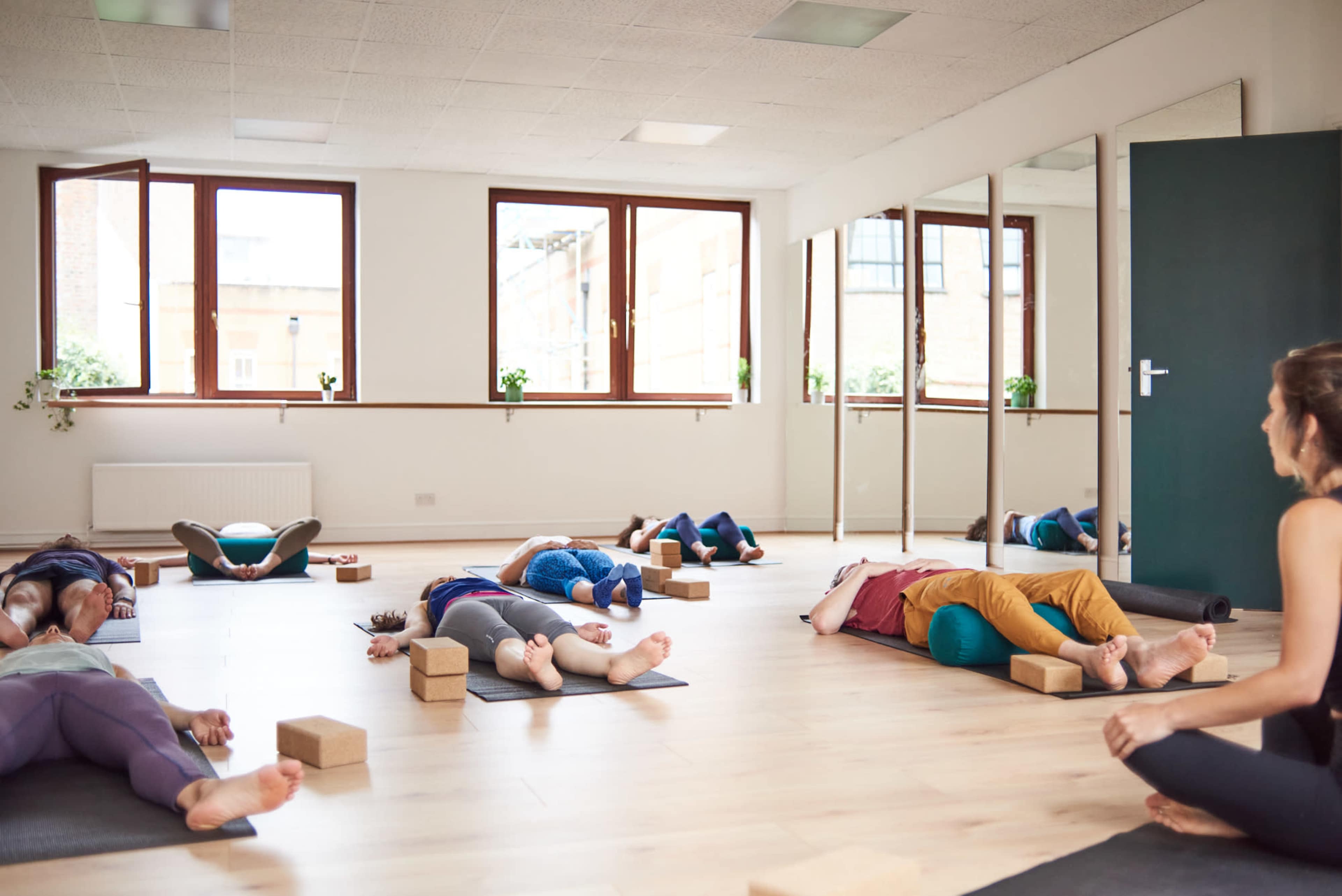 A group of people are practicing yoga in a bright studio, lying on mats with blocks for support.
