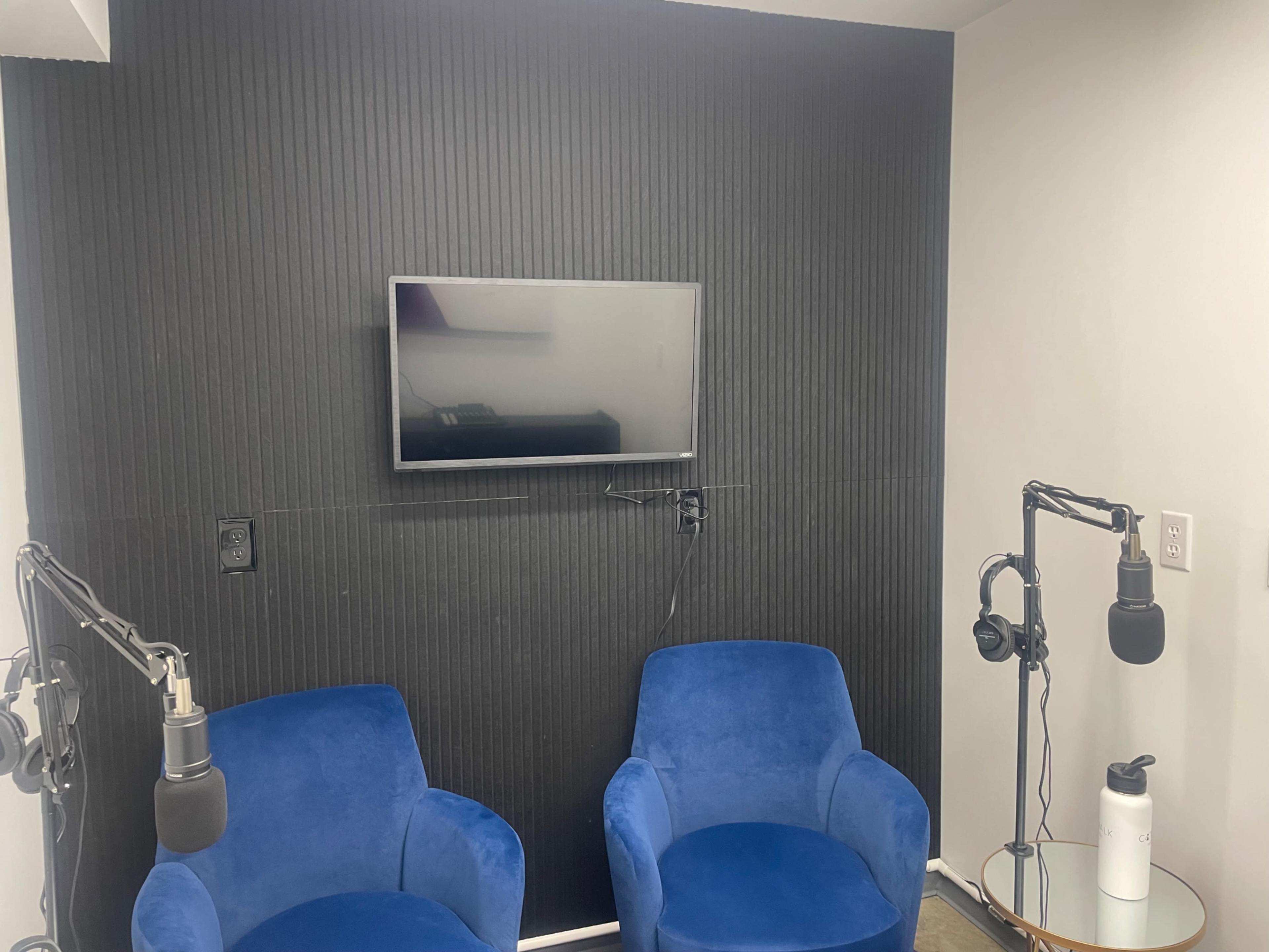 The image shows a small recording studio with two blue chairs, a television mounted on a black ribbed wall, and microphone stands positioned for a podcast setup.