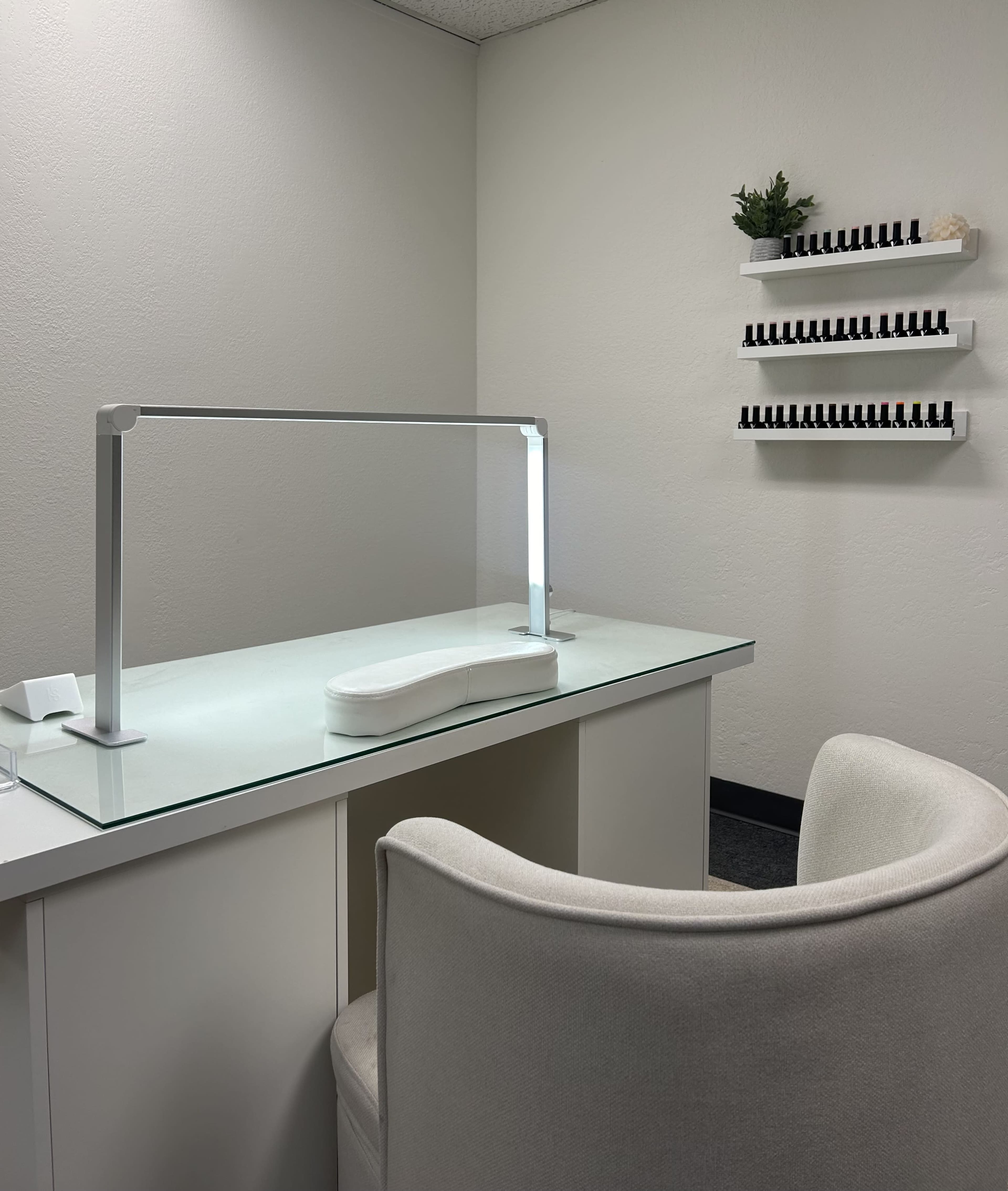 A minimalist nail salon features a glass-topped desk with a lamp and a white chair, alongside a shelf displaying various nail polish bottles.