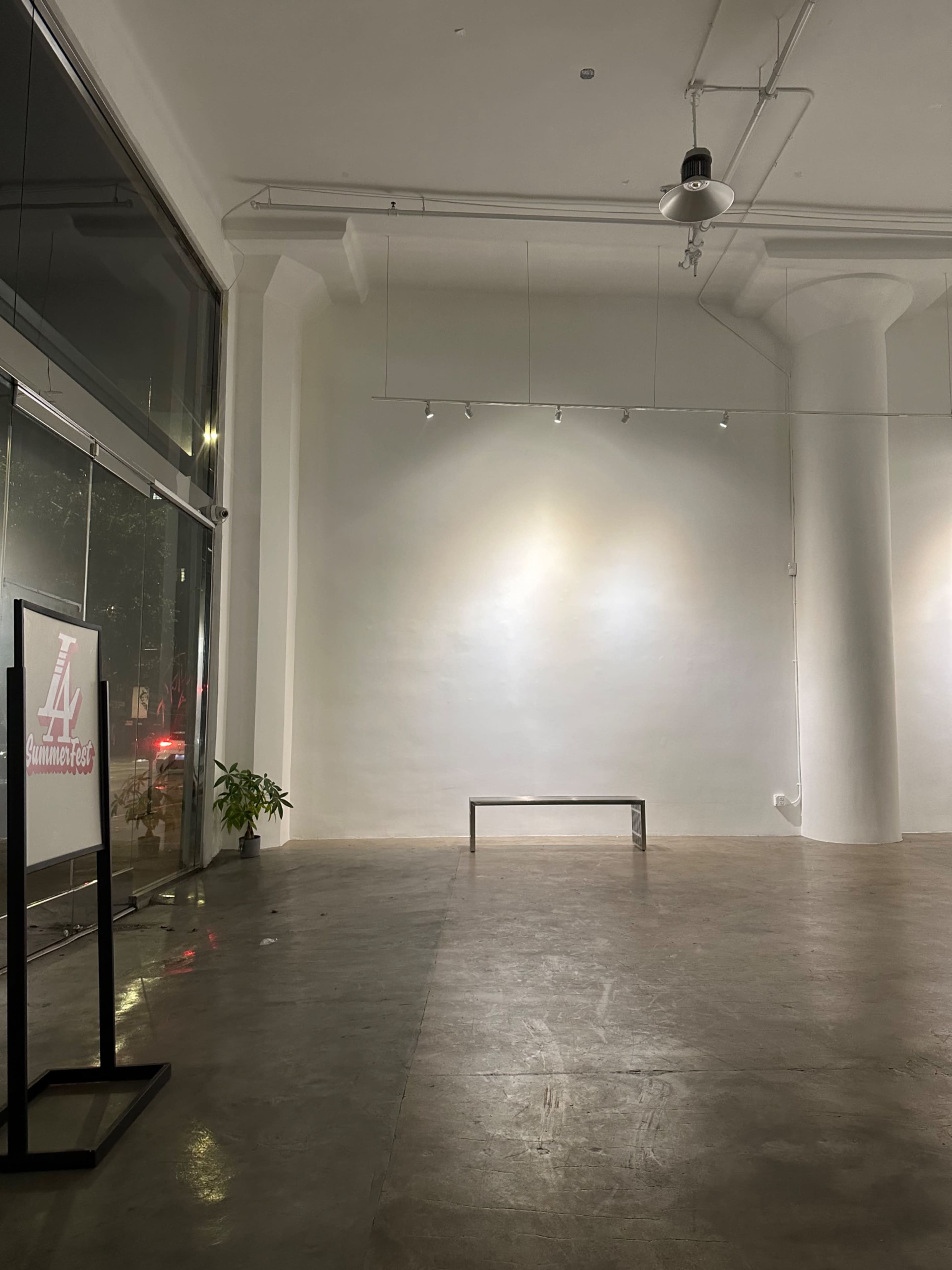 The image shows a minimalist room with a blank white wall, a wooden bench in the center, and a small potted plant to the left, illuminated by soft lighting.