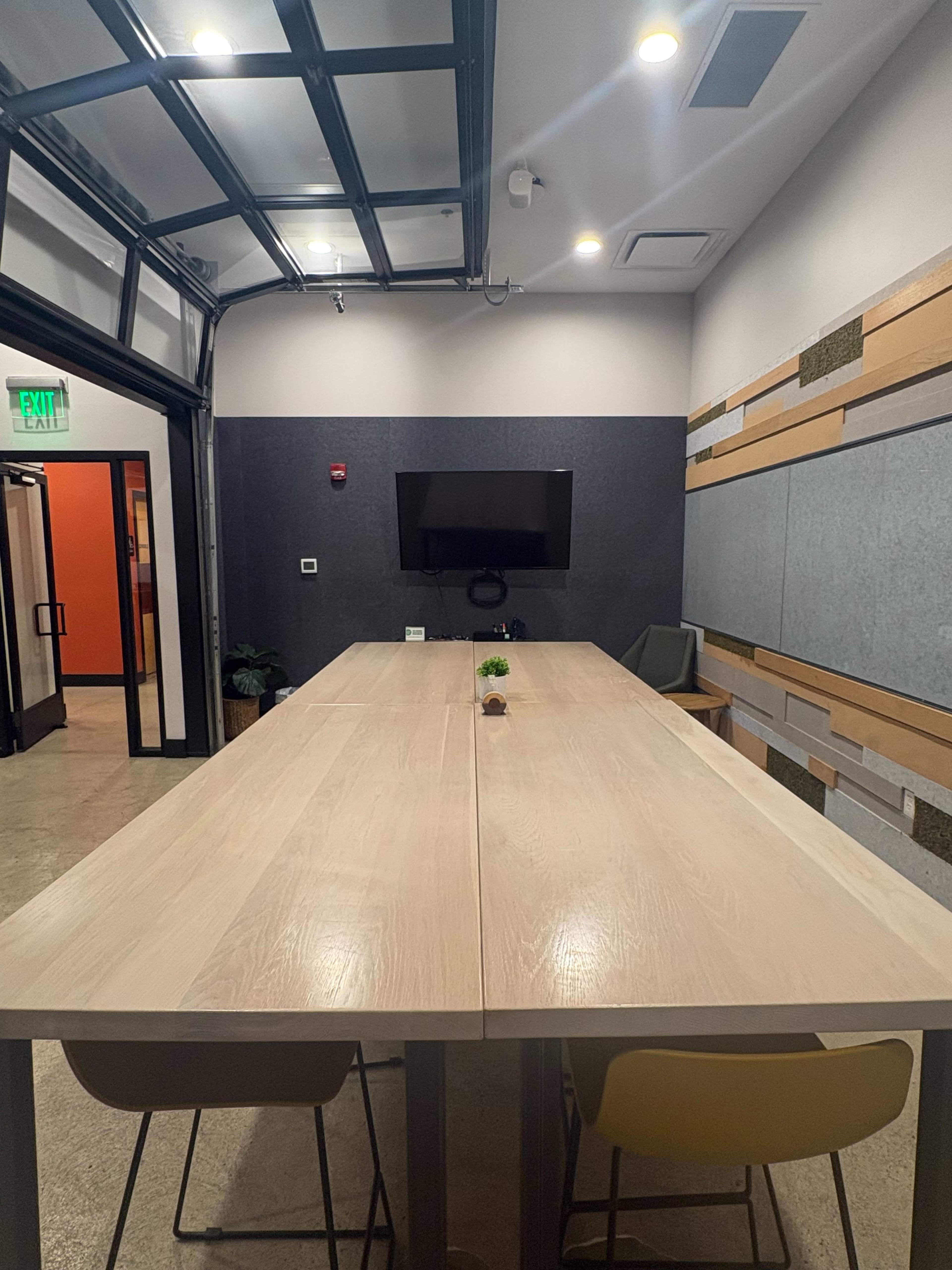 The image shows a modern conference room with a long wooden table, a television mounted on the wall, and large windows with glass doors.