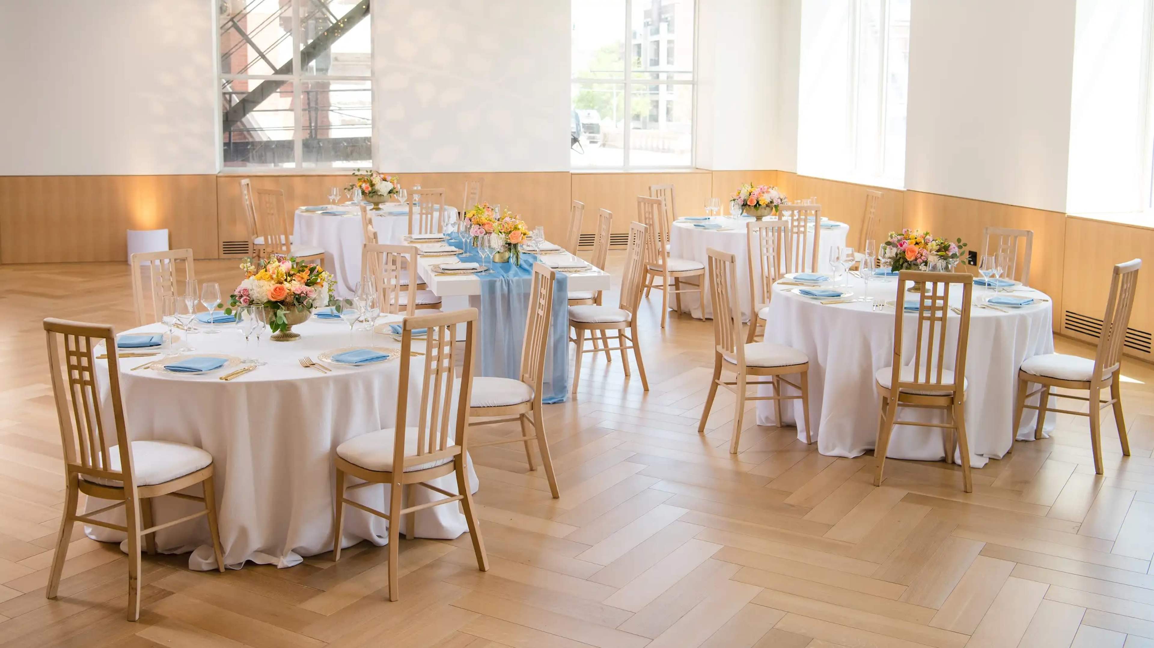 The image shows a banquet hall arranged with several round tables covered in white tablecloths, each adorned with floral centerpieces and place settings, and bathed in natural light from large windows.