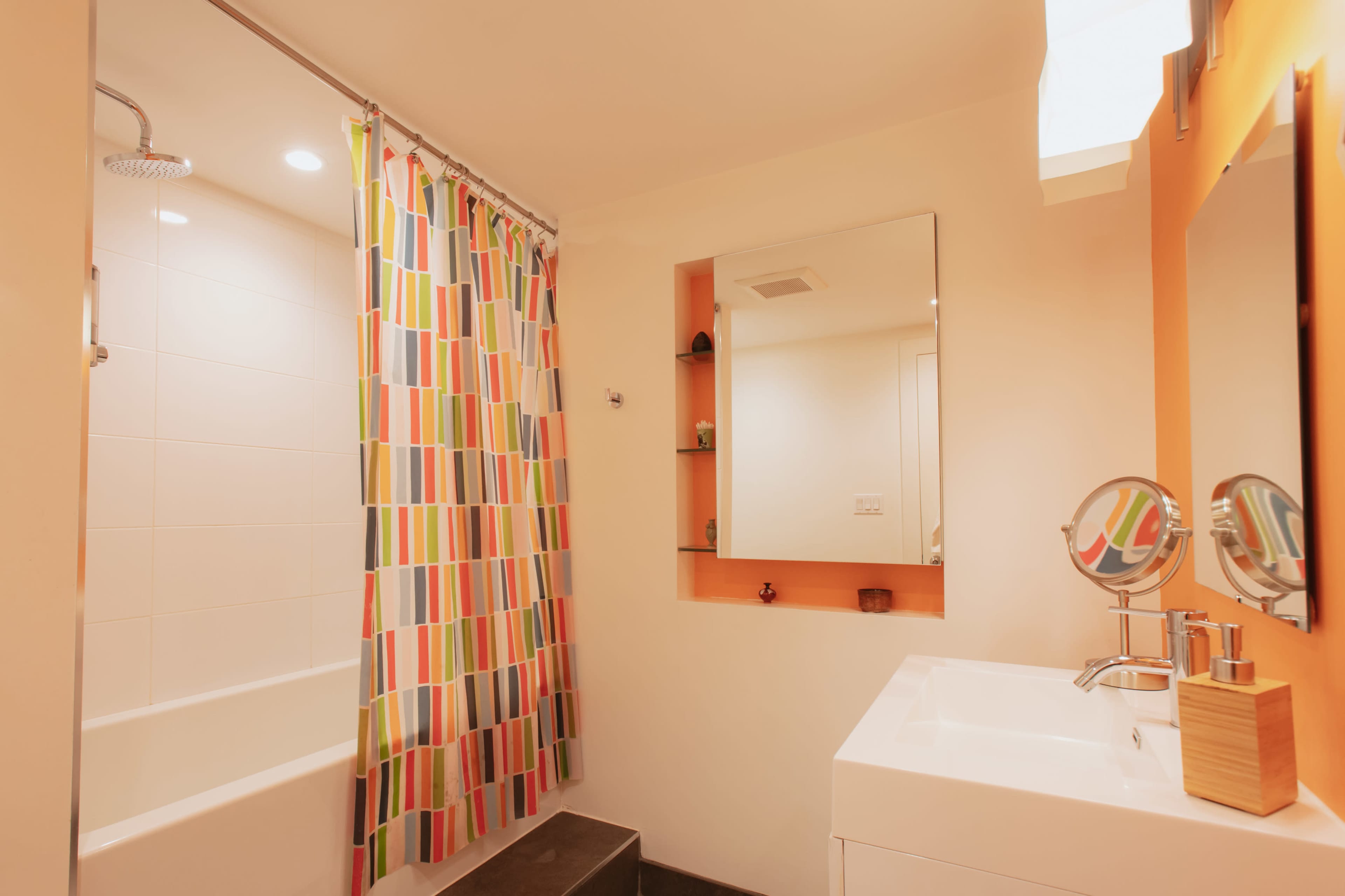 The bathroom features a shower with a colorful striped curtain, a rectangular mirror above a sink, and a wall-mounted shelf.