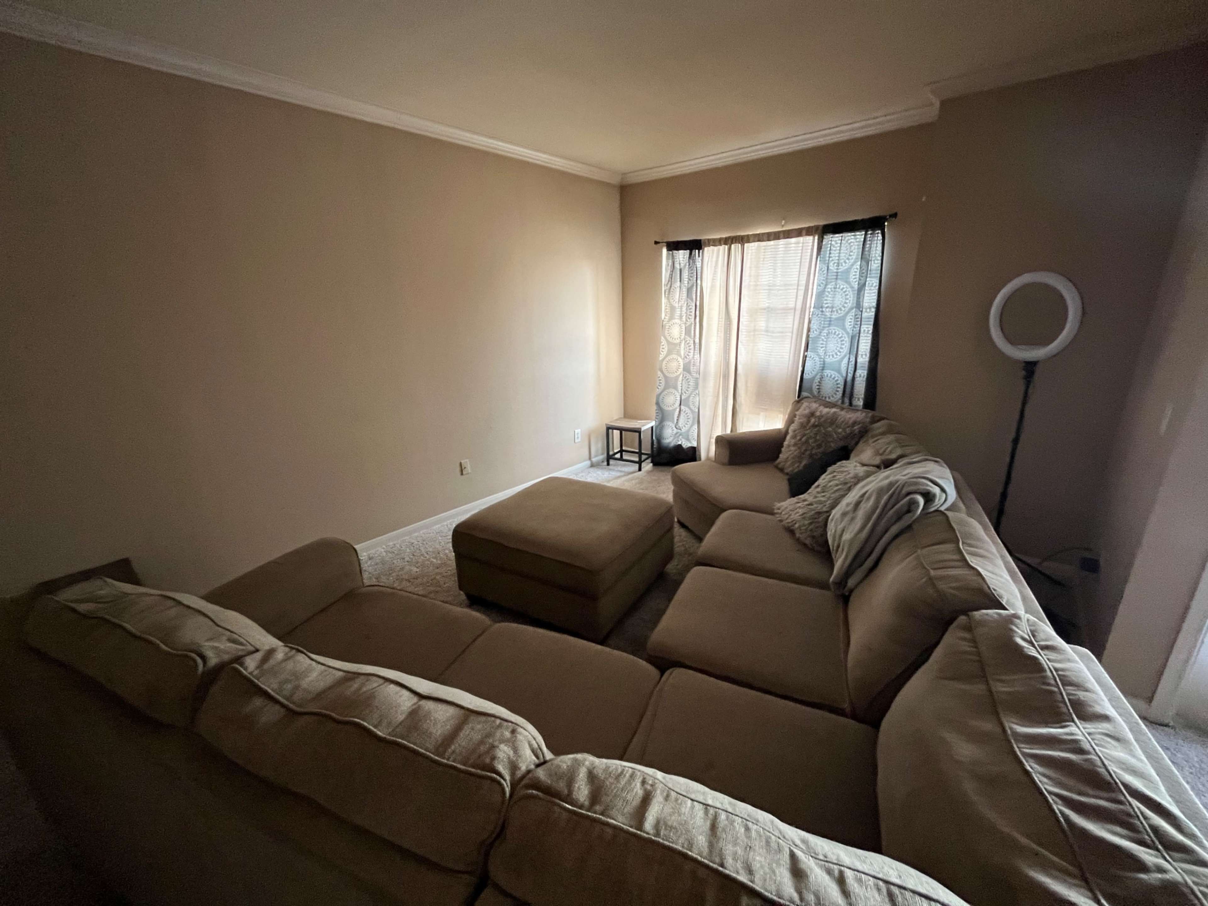 A spacious living room with a large sectional sofa, a small table, and a ring light next to a window with sheer curtains.