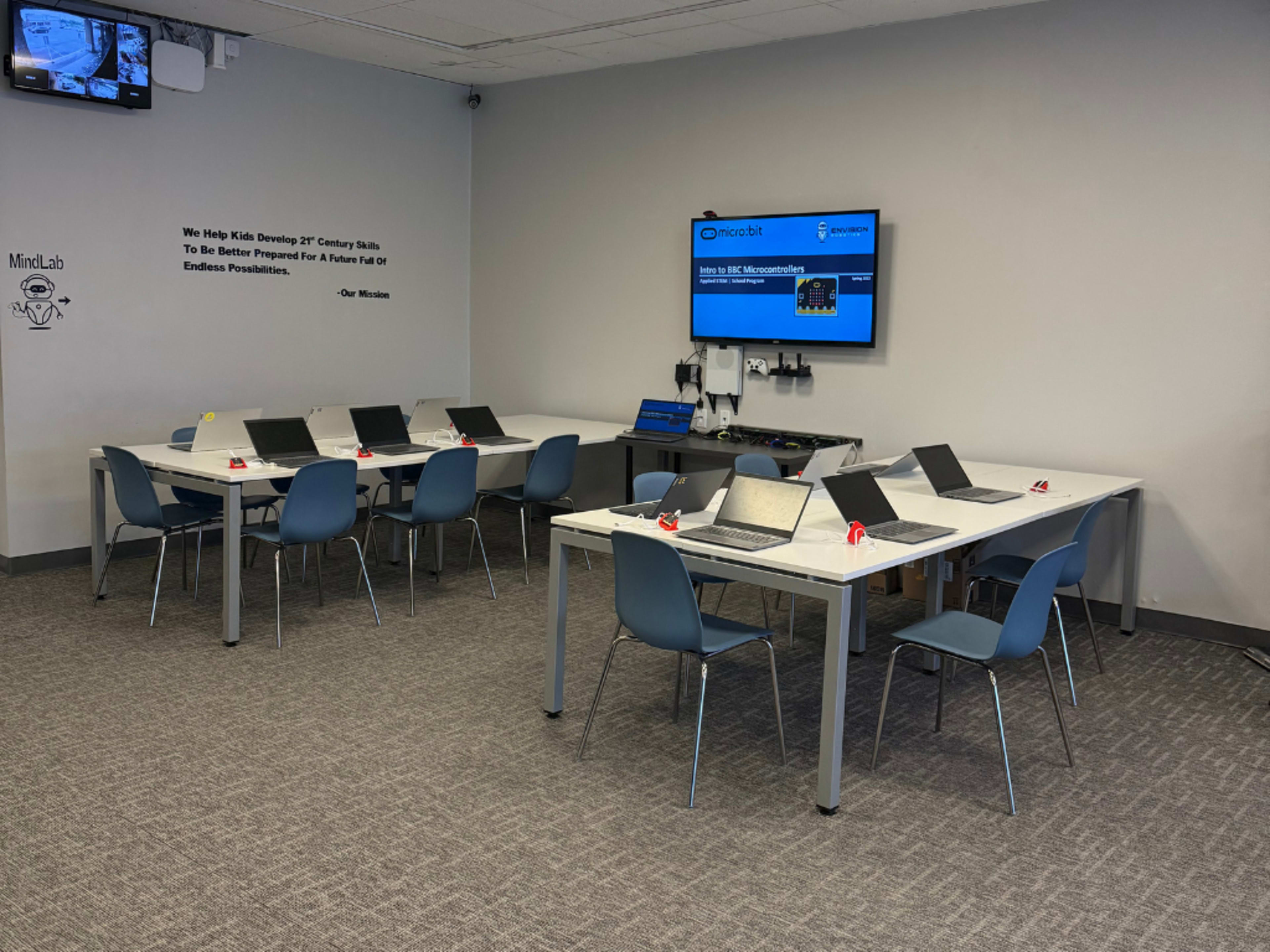 The image shows a modern classroom with multiple tables arranged in a U-shape, each equipped with laptops and chairs, along with a large screen displaying educational content.