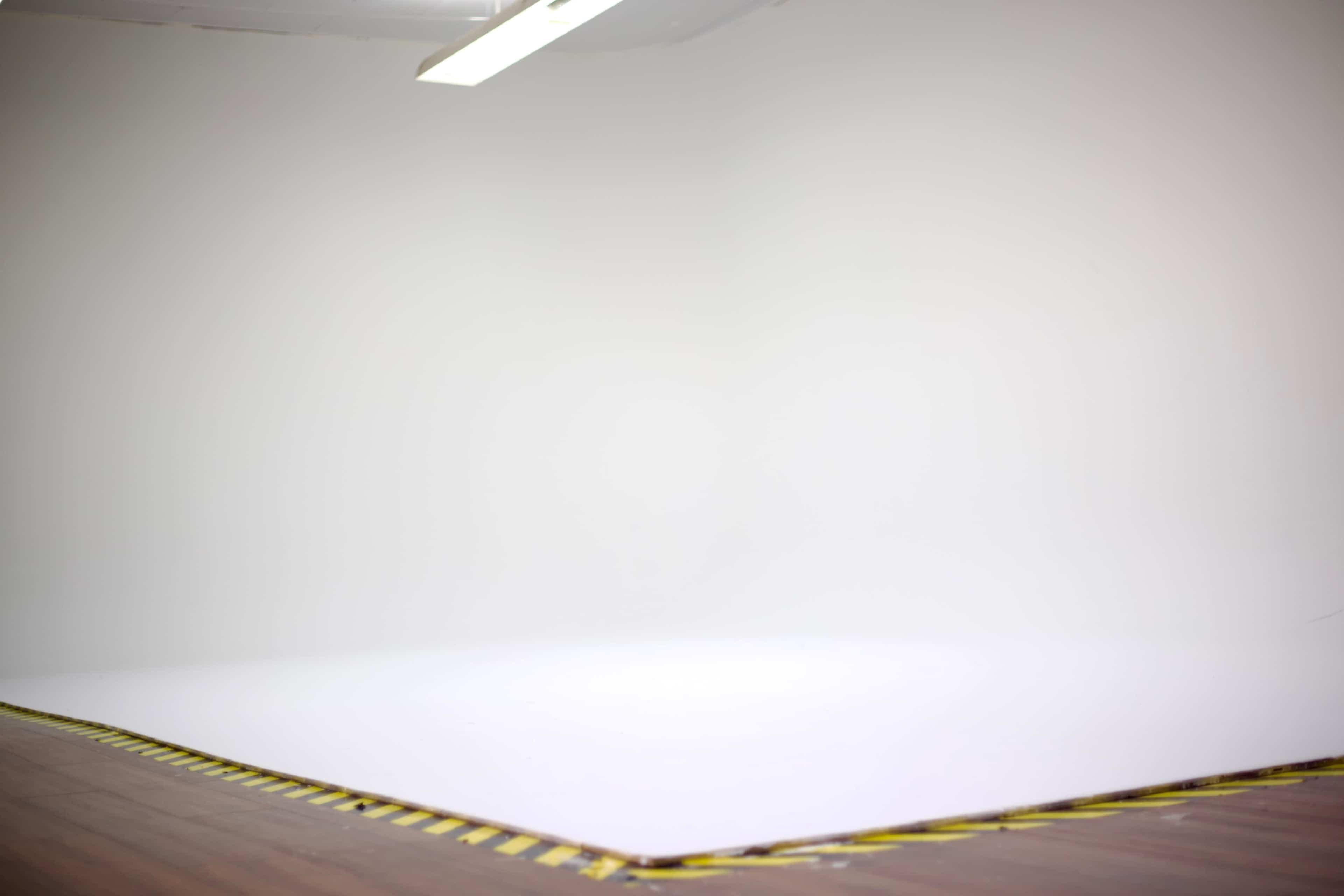 The image shows a minimalistic studio space with a white backdrop and wooden flooring, bordered by caution tape.