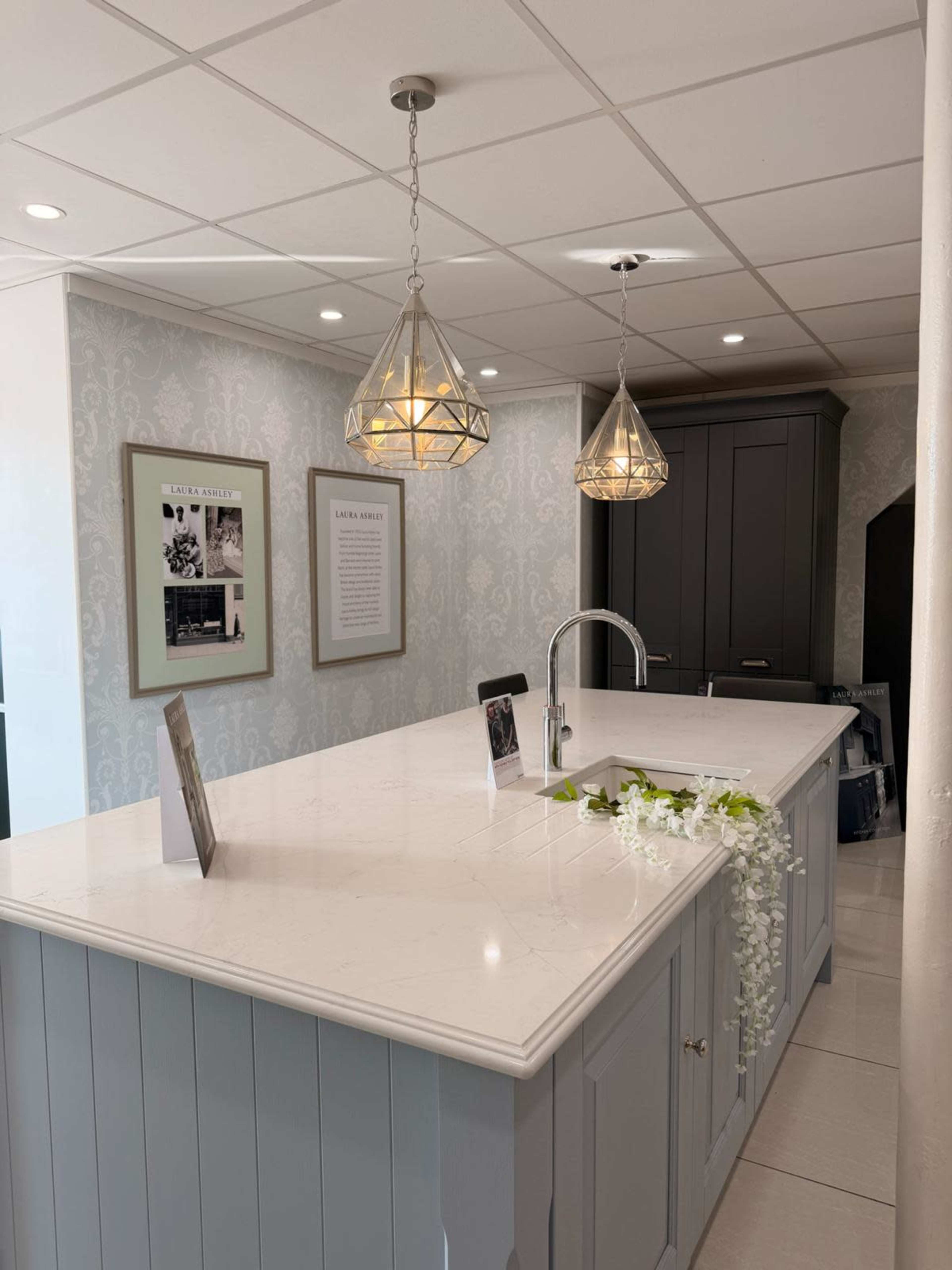 The image shows a modern kitchen with a light gray island, pendant lights hanging above it, and decorative wall features.