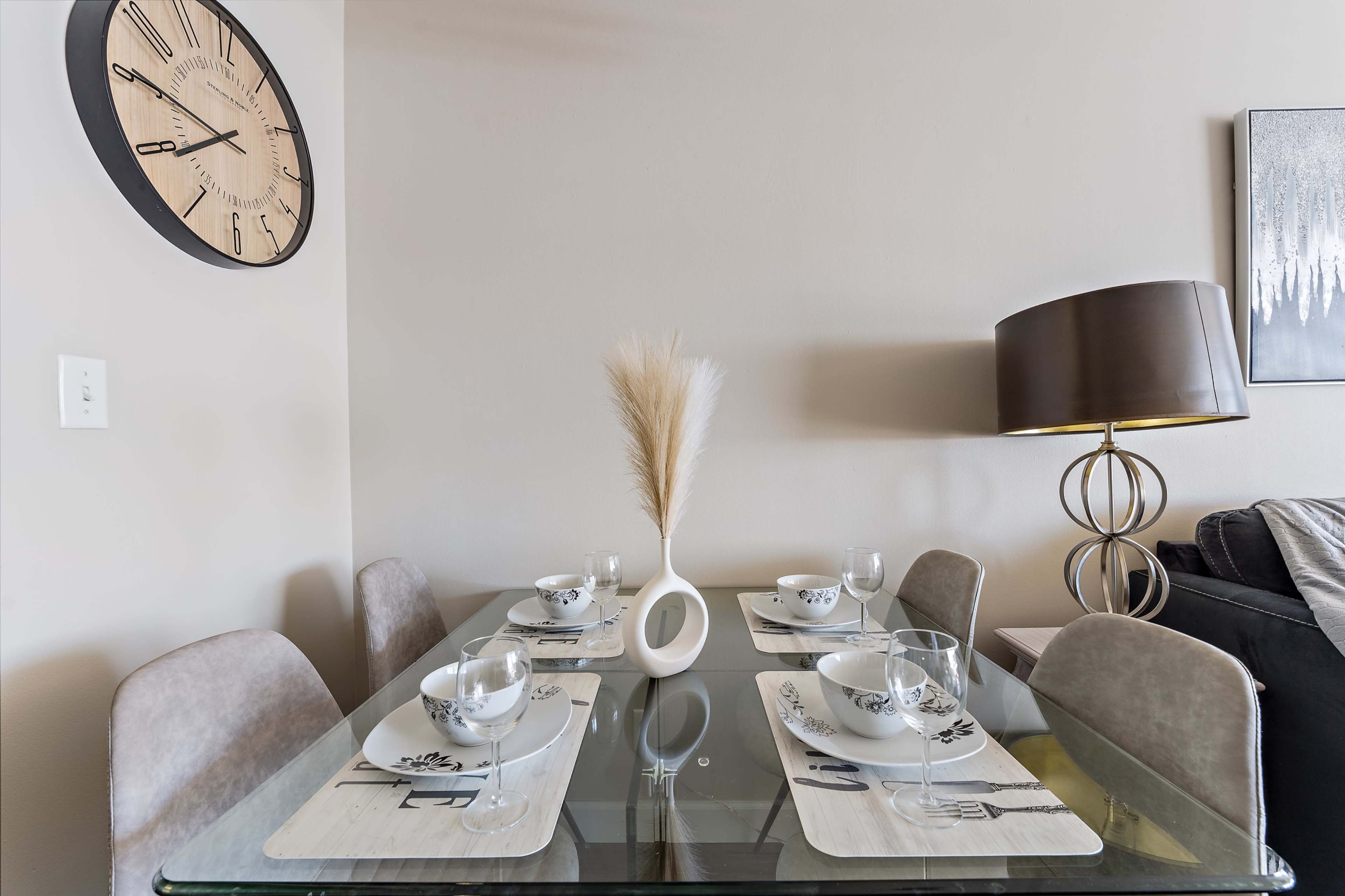 A modern dining table is elegantly set with six bowls, placemats, and a decorative vase with a plume, accompanied by a wall clock and a stylish lamp in the background.