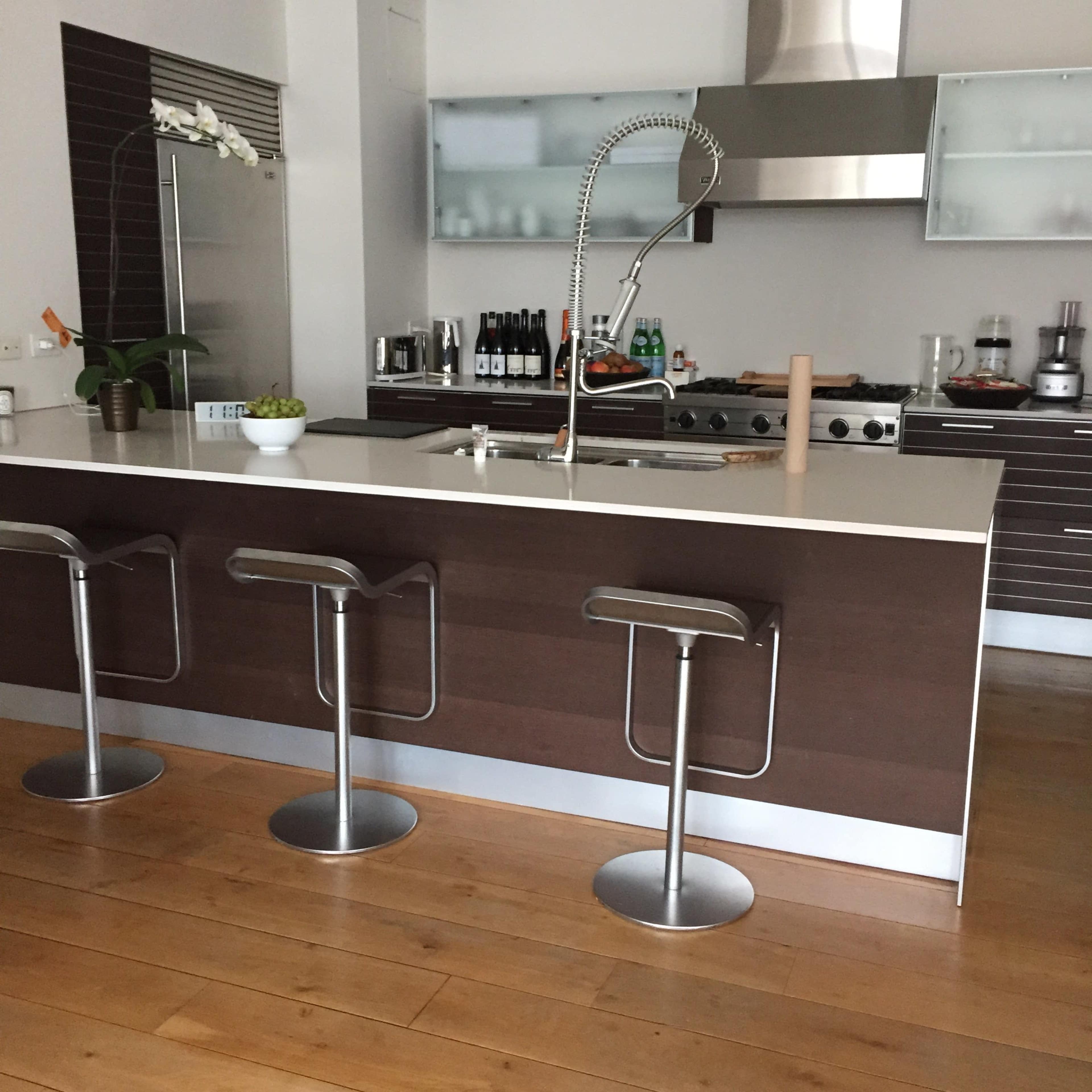 The image shows a modern kitchen with a central island, featuring three barstools, stainless steel appliances, and a sleek countertop.
