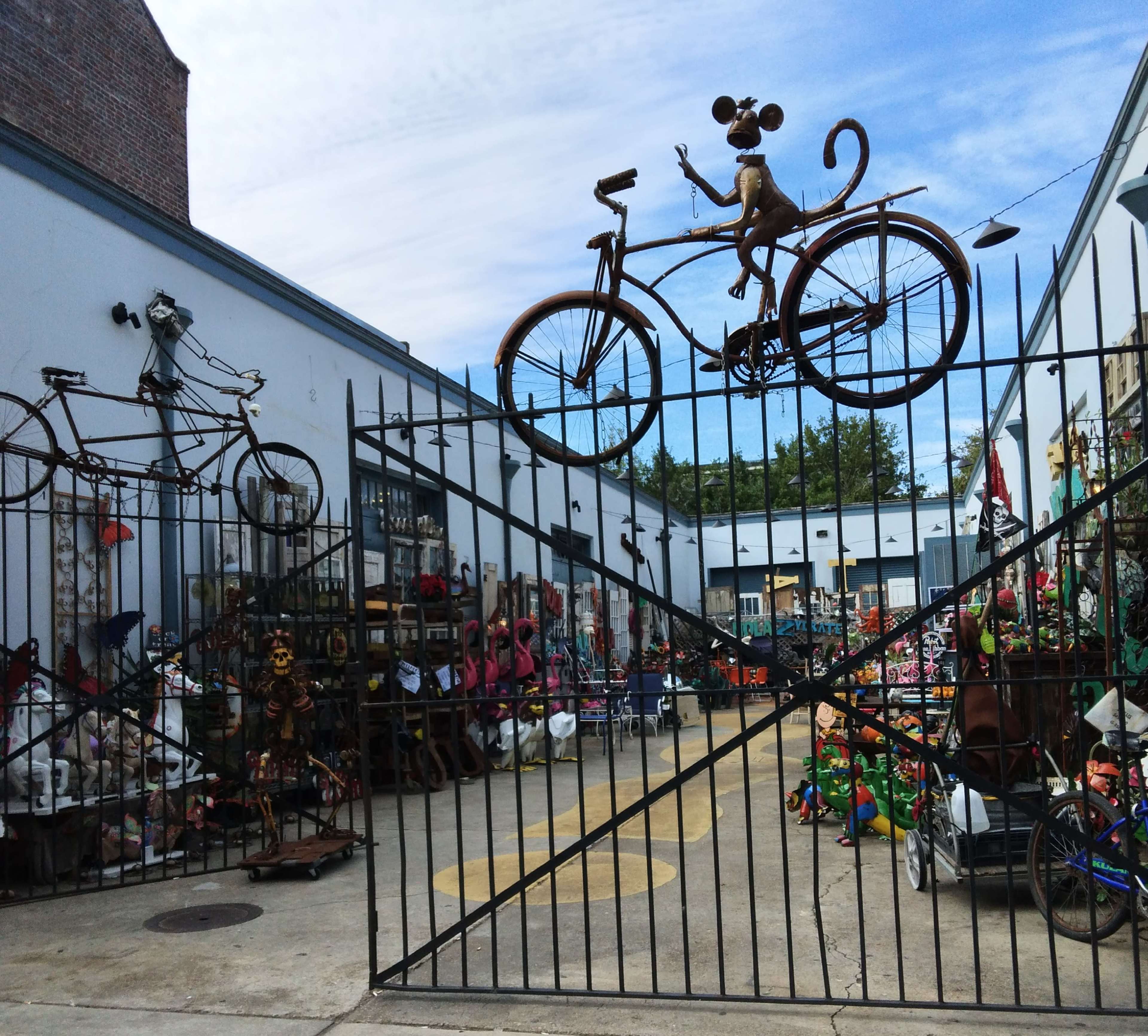 The image shows a decorative metal gate with a bicycle and a monkey sculpture on top, leading into a vibrant outdoor space filled with various sculptures and items for sale.