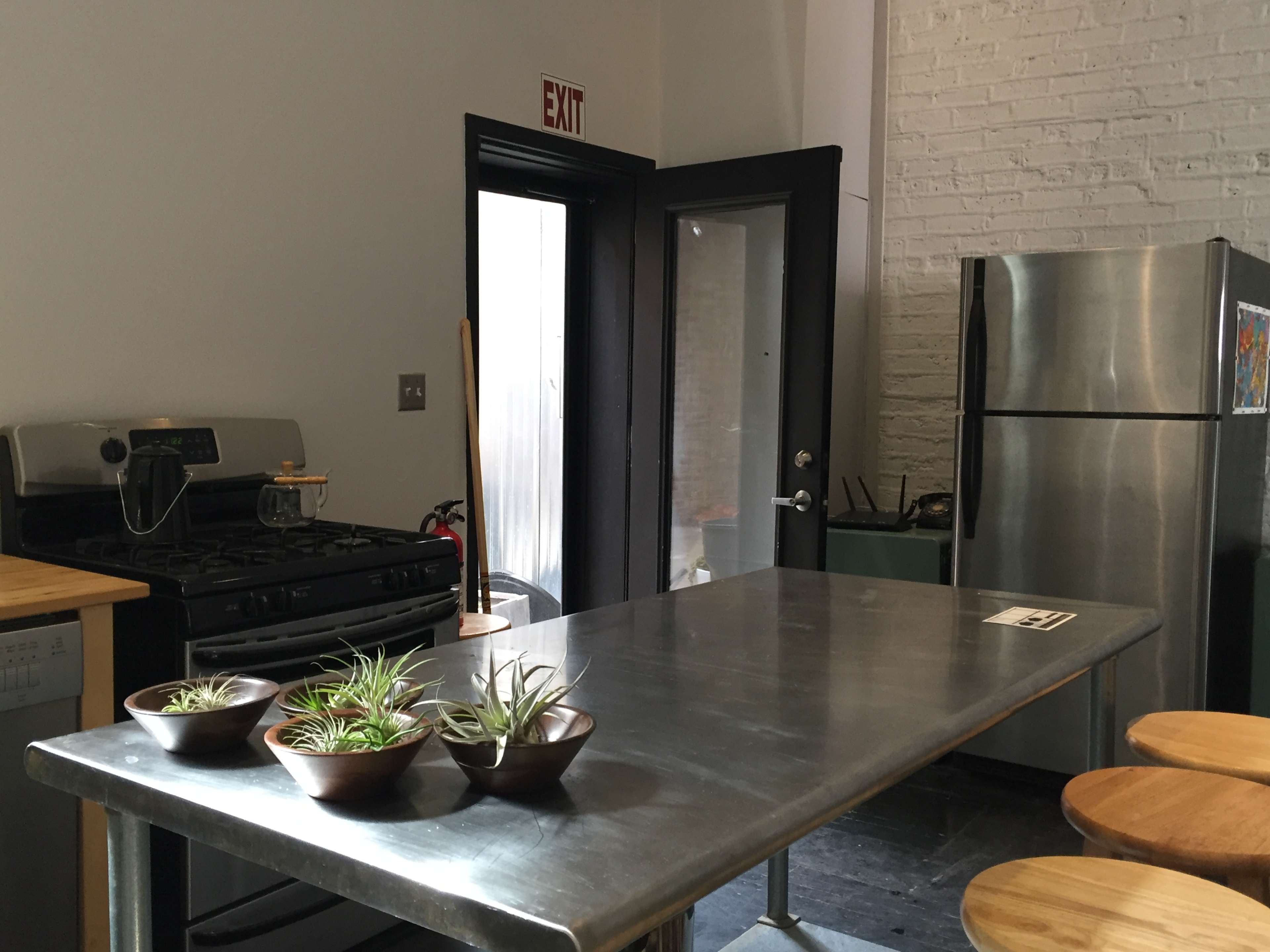 A kitchen with a stainless steel table, several bowls of plants, a gas stove, a refrigerator, and an exit door.