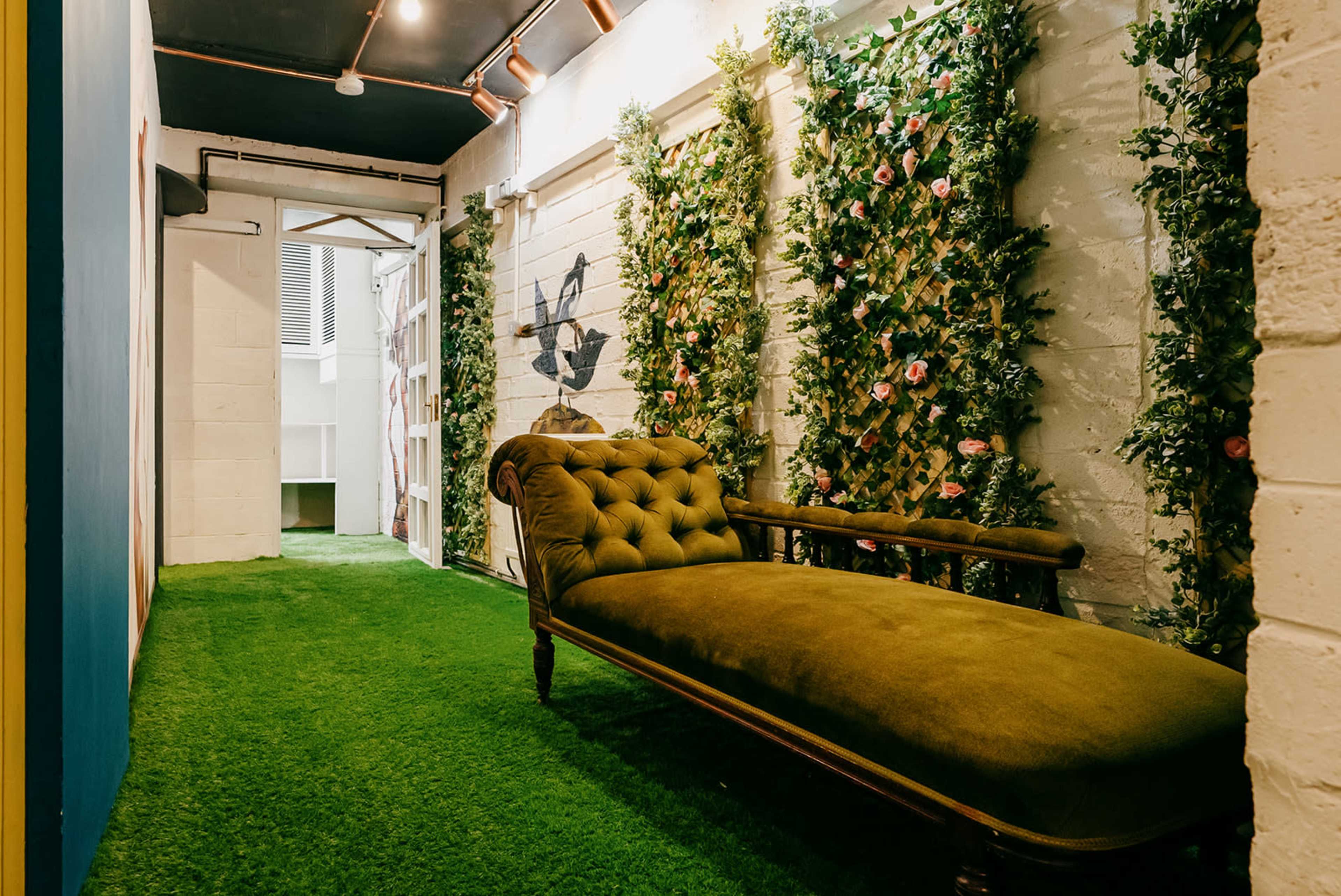 A hallway with artificial grass flooring, flanked by green walls adorned with plants and flowers, leading to an open door.