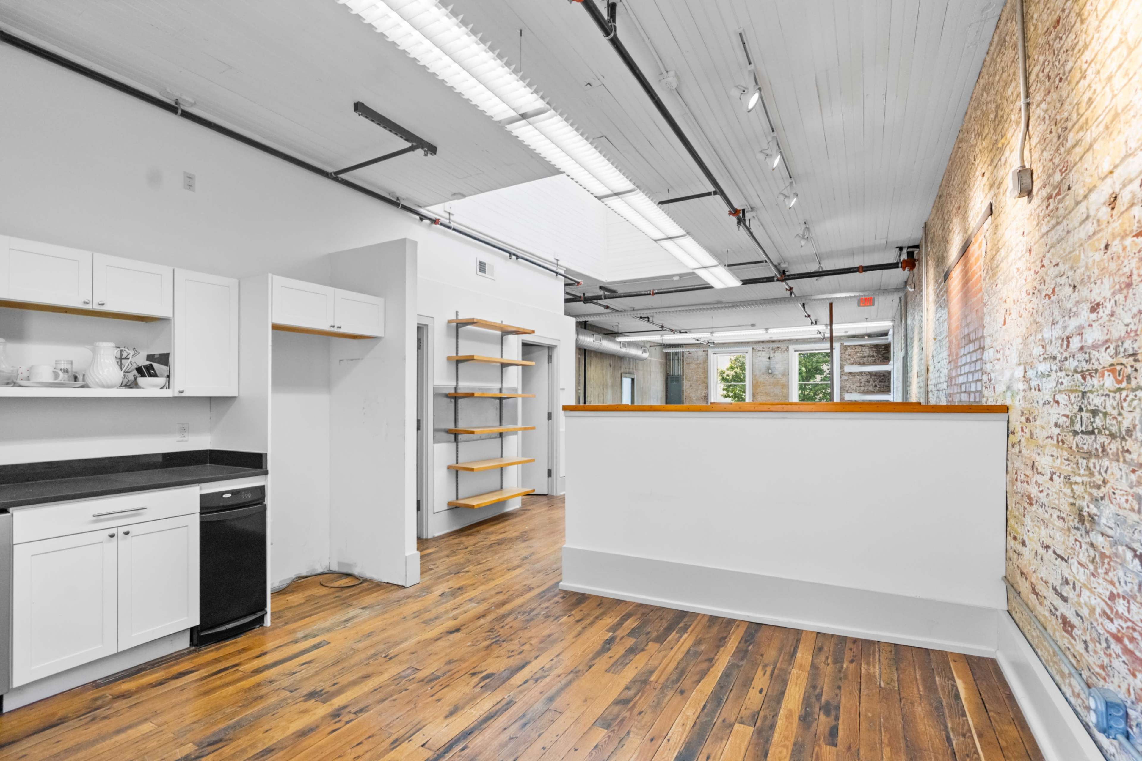 The image shows a spacious room with wooden flooring, a kitchen area featuring white cabinets and a dishwasher, and a brick wall on one side.
