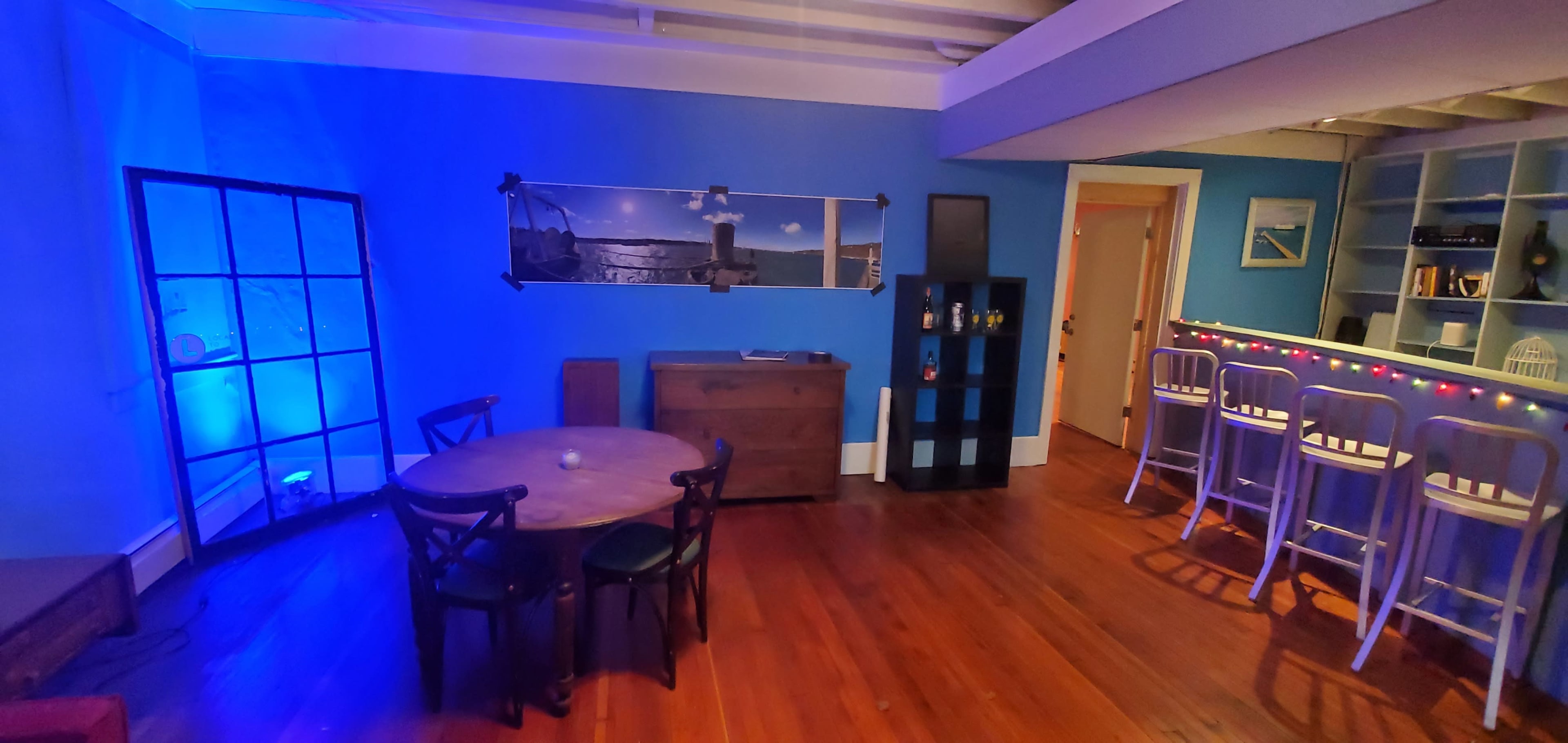 A room featuring a circular wooden table with four chairs, a blue wall with a large photograph, a sideboard, and a bar with high stools.