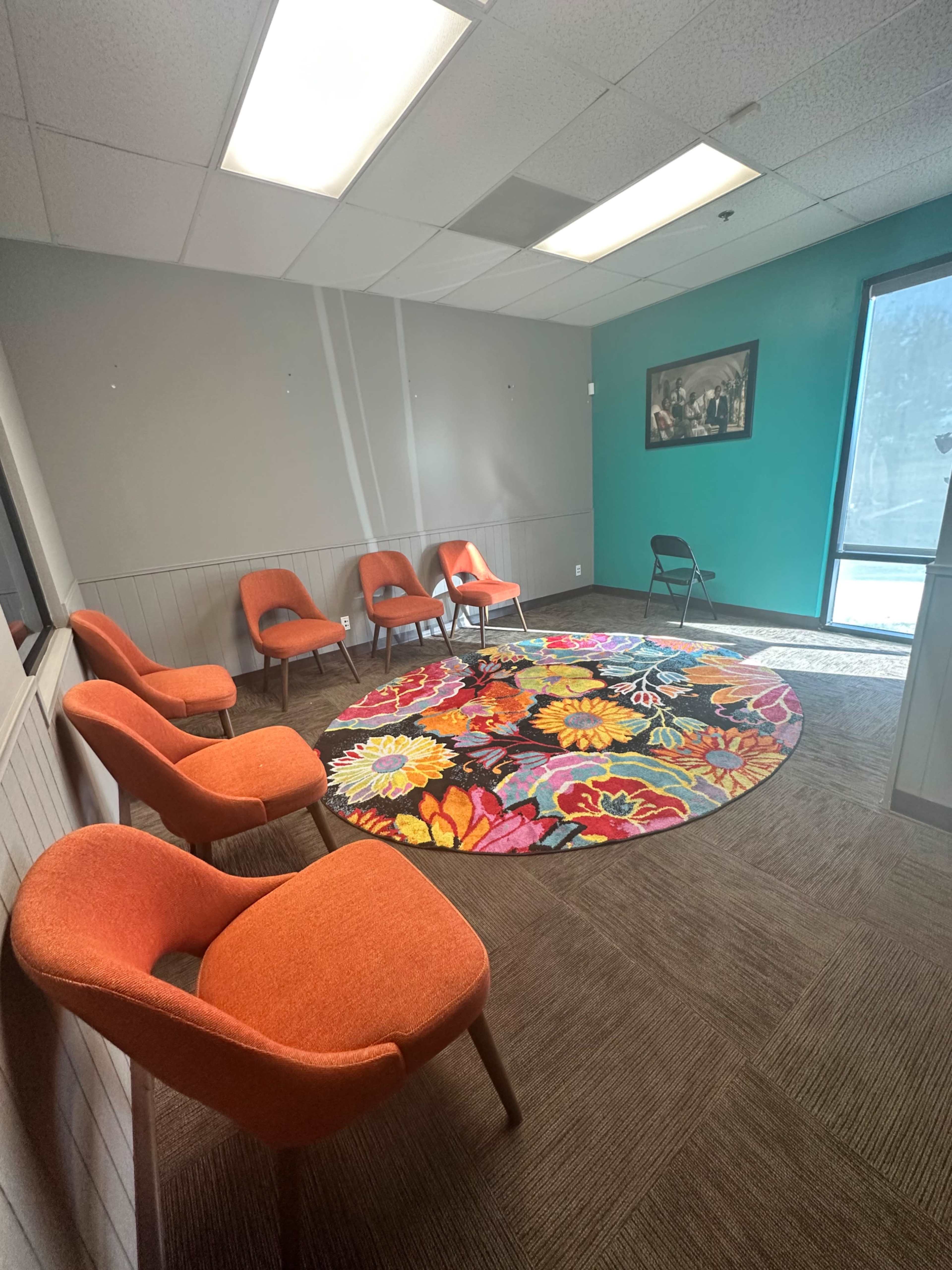 A waiting area with five orange chairs arranged around a colorful floral rug, complemented by a teal accent wall and a large window letting in natural light.
