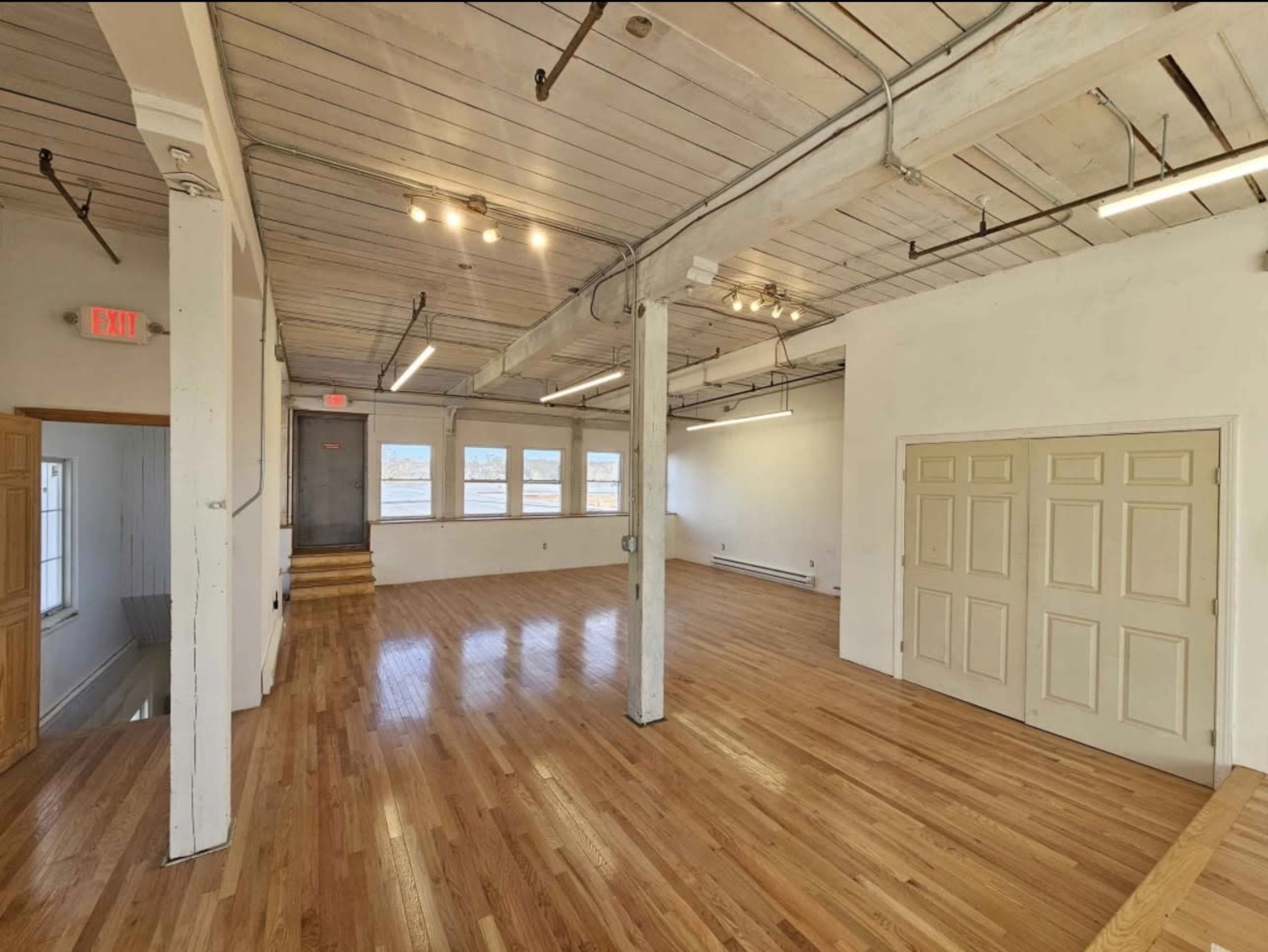The image shows a spacious, empty room with polished wooden floors, white walls, and large windows, illuminated by overhead lights.