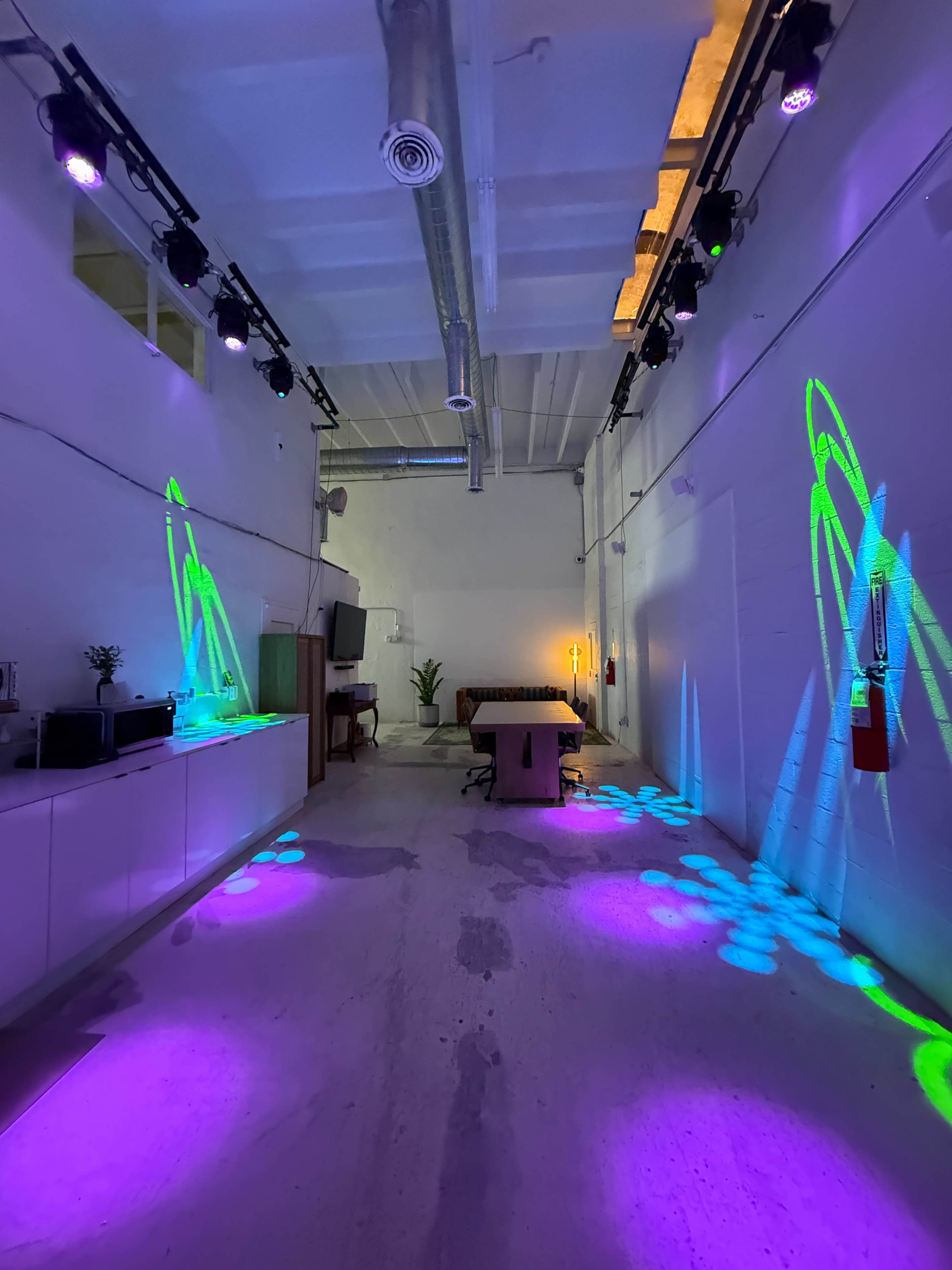 The image shows a spacious room with colorful lights projecting patterns on the floor and walls, featuring a large table in the center and a plant in the background.