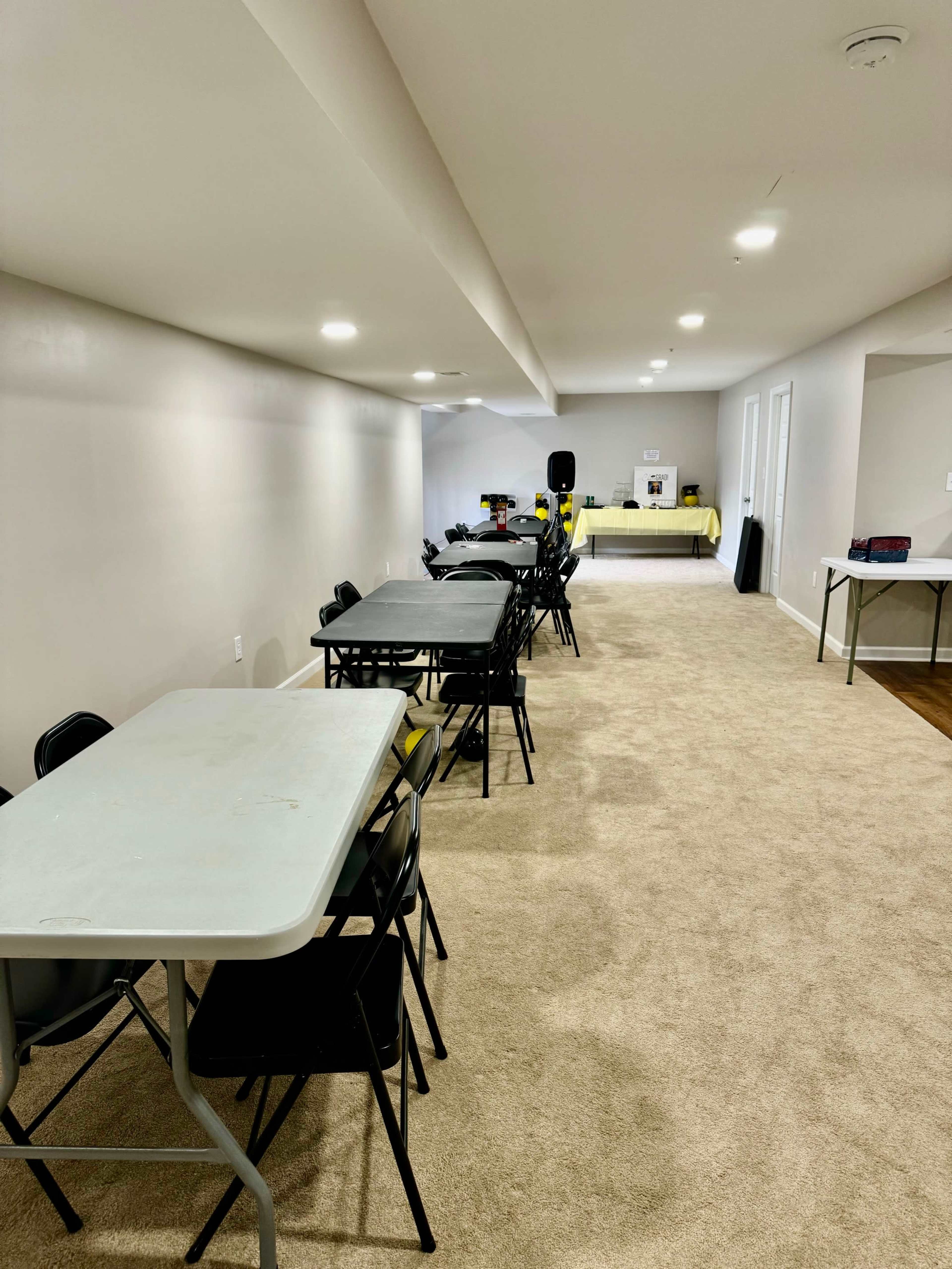 A long, carpeted room features several rows of folding tables and chairs, with a stage area at the far end.