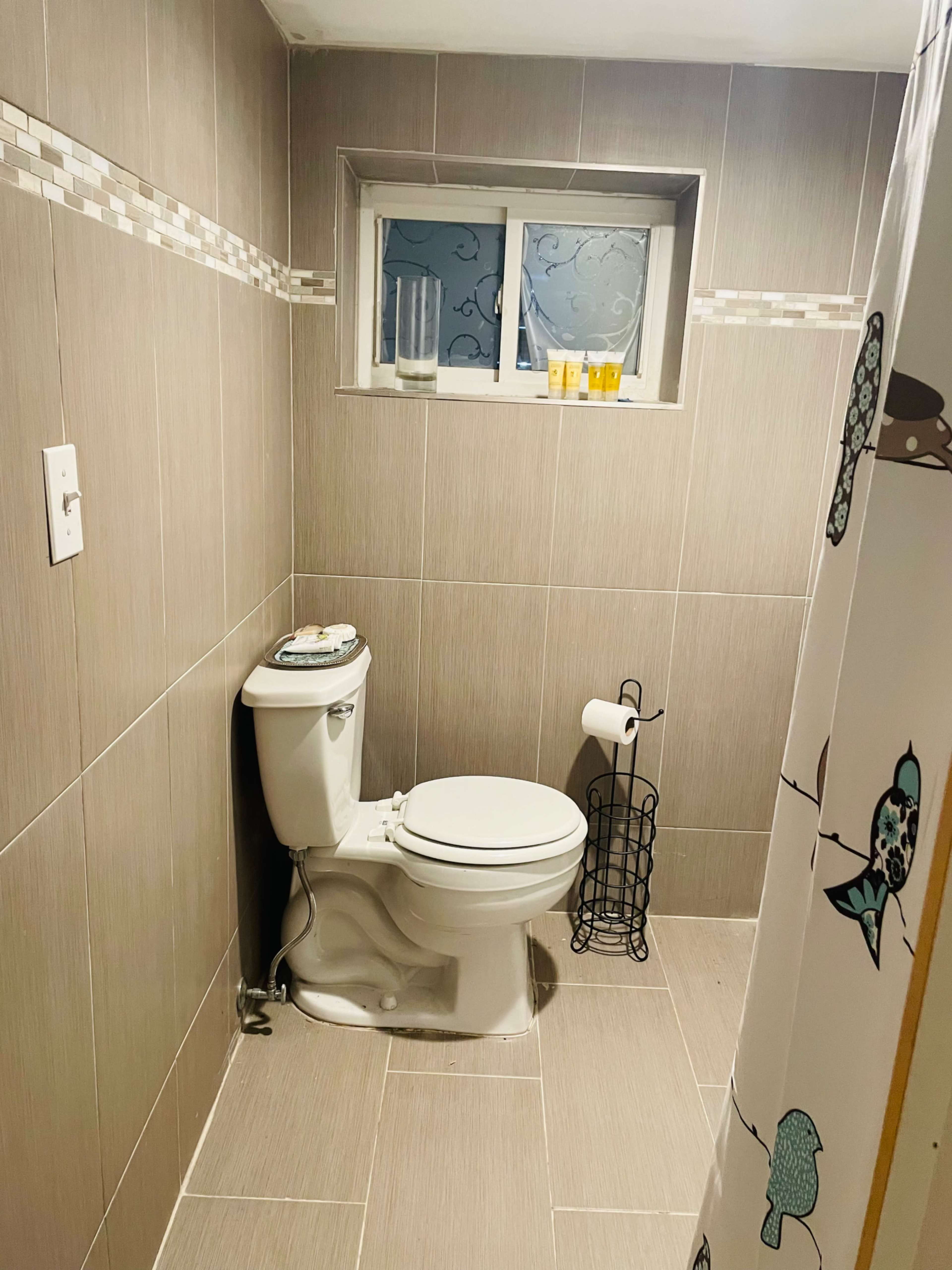 The image shows a modern bathroom with beige tiled walls, a toilet, and a window above the toilet.