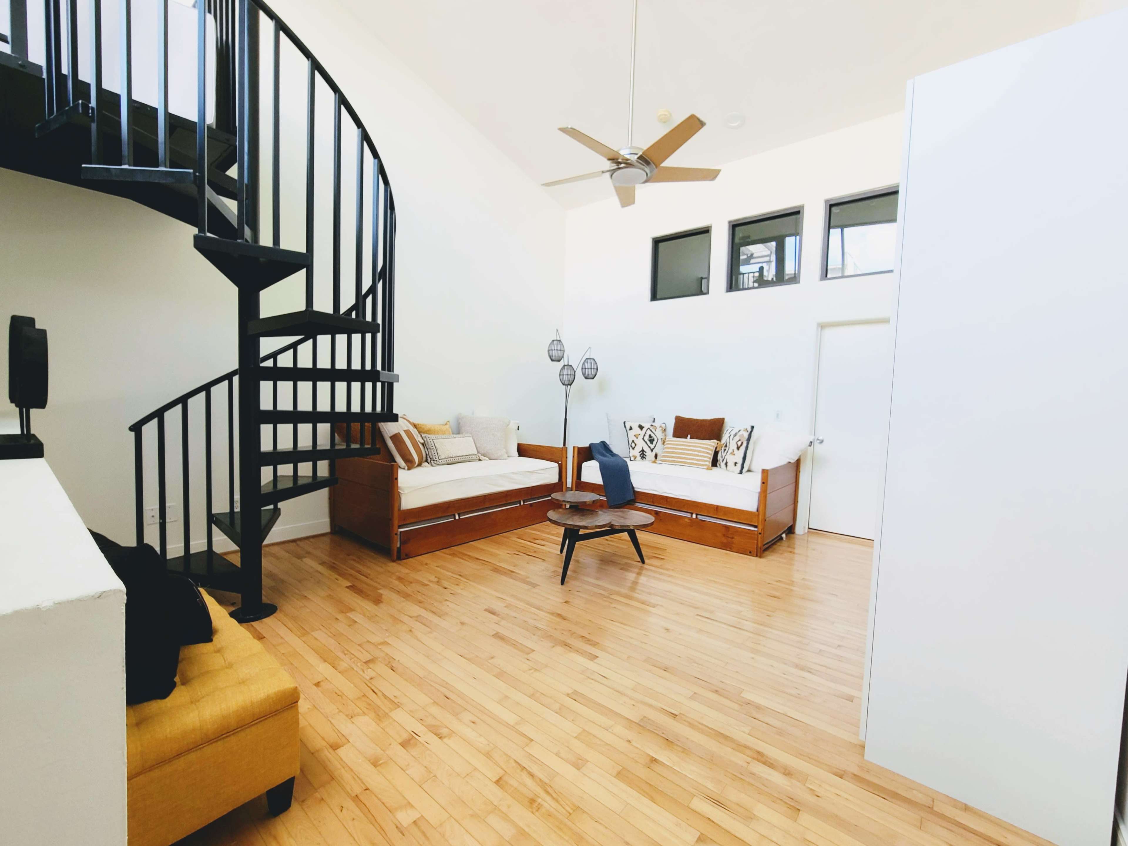 A spacious room with wooden flooring, a spiral staircase, two daybeds, and a ceiling fan.