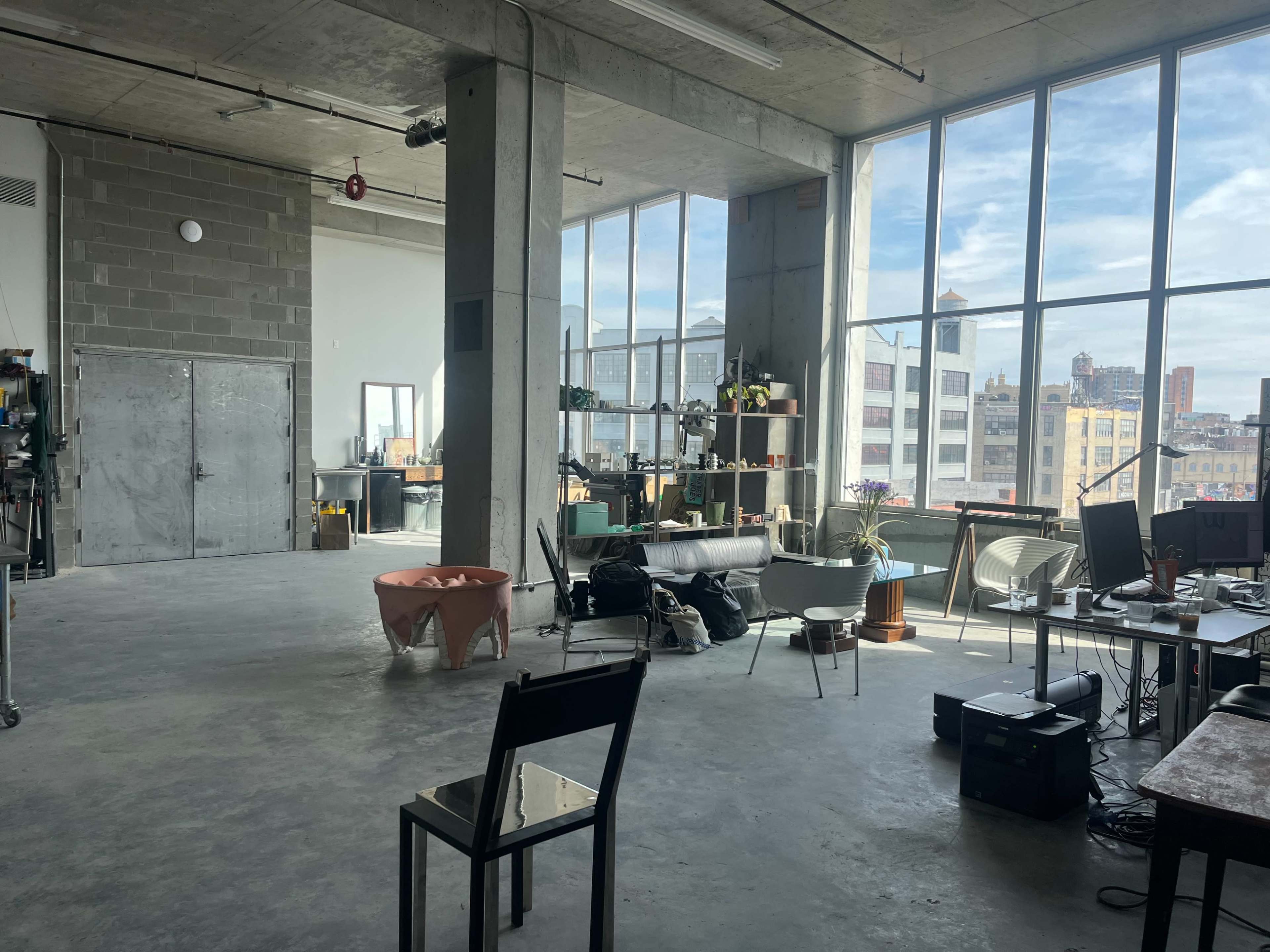 The image shows a spacious industrial-style room with large windows, concrete walls, and a mix of furniture, including a chair, a couch, and several workstations.