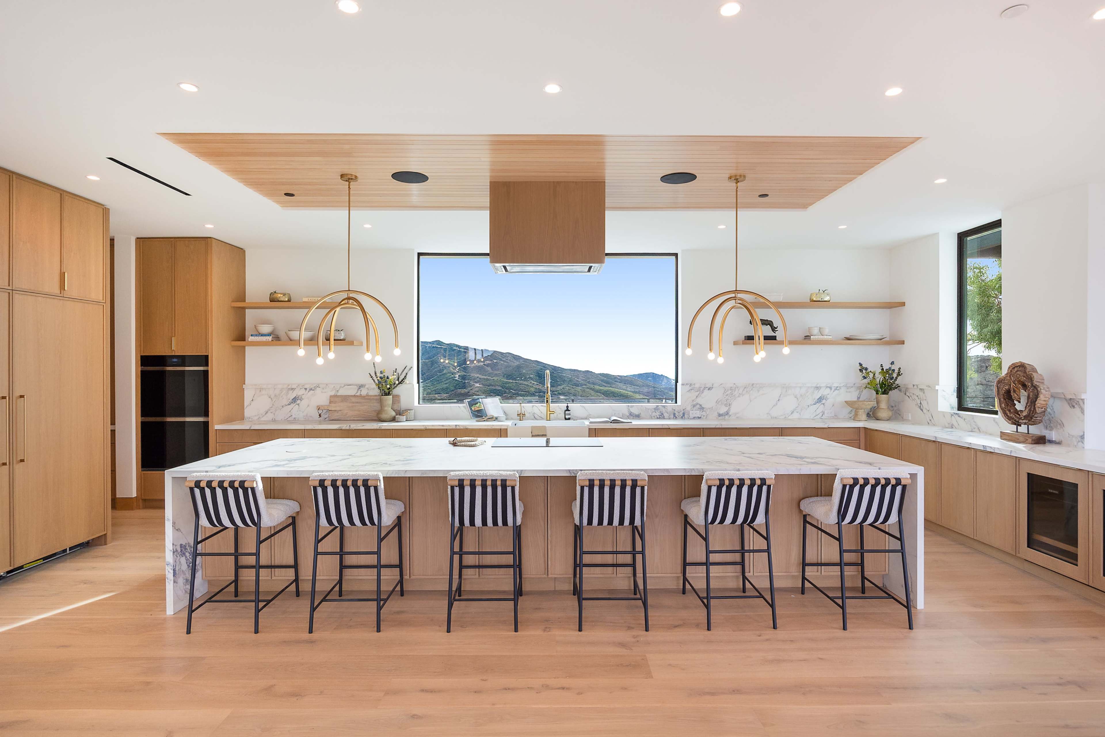 The kitchen features a large island with seating, wooden cabinetry, and a panoramic window showcasing a mountain view.