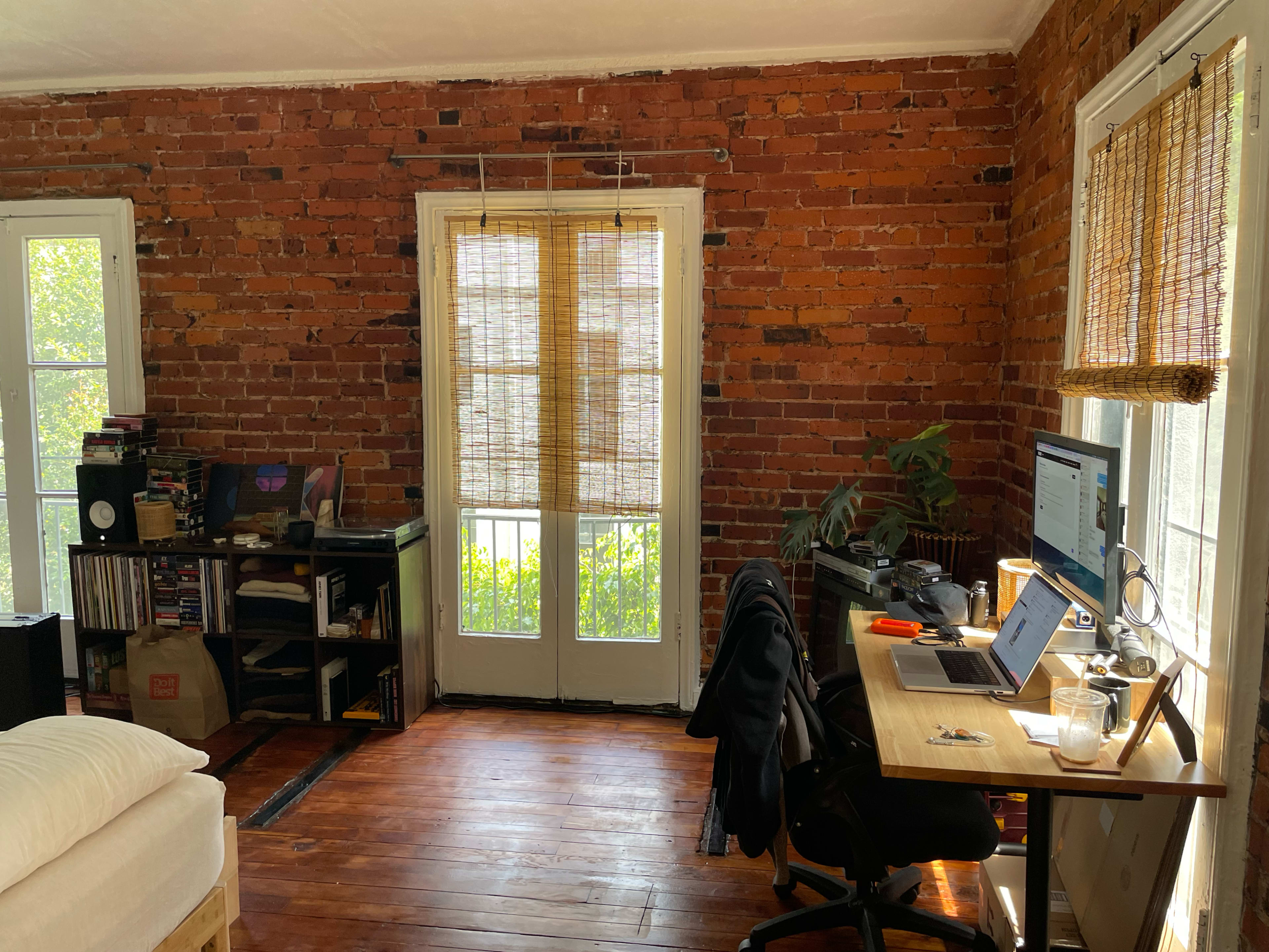 The image shows a cozy room with exposed brick walls, a desk with a computer and various office supplies, and large windows covered by bamboo shades allowing natural light to enter.