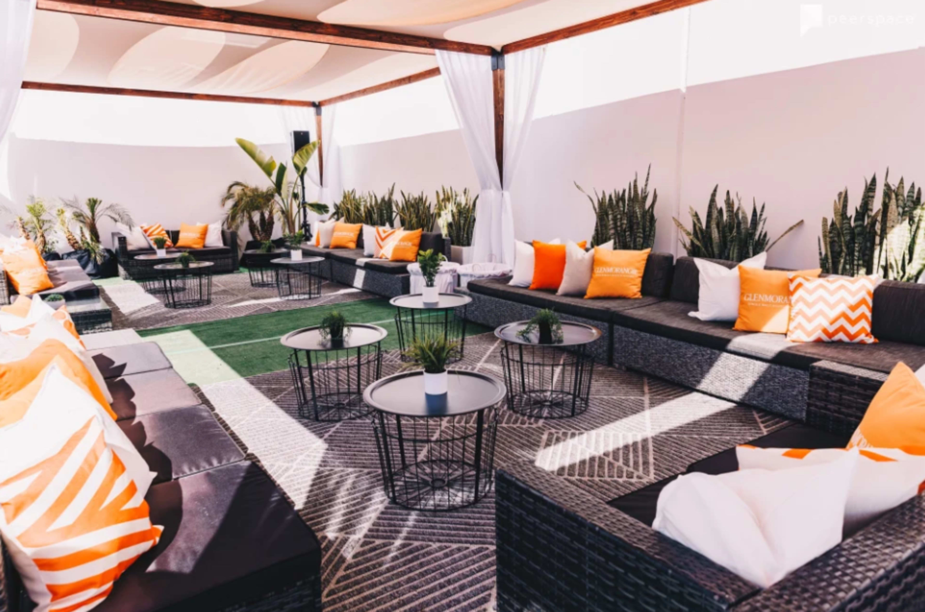 A modern outdoor lounge area features black wicker seating with orange and white cushions, low tables, and decorative plants.
