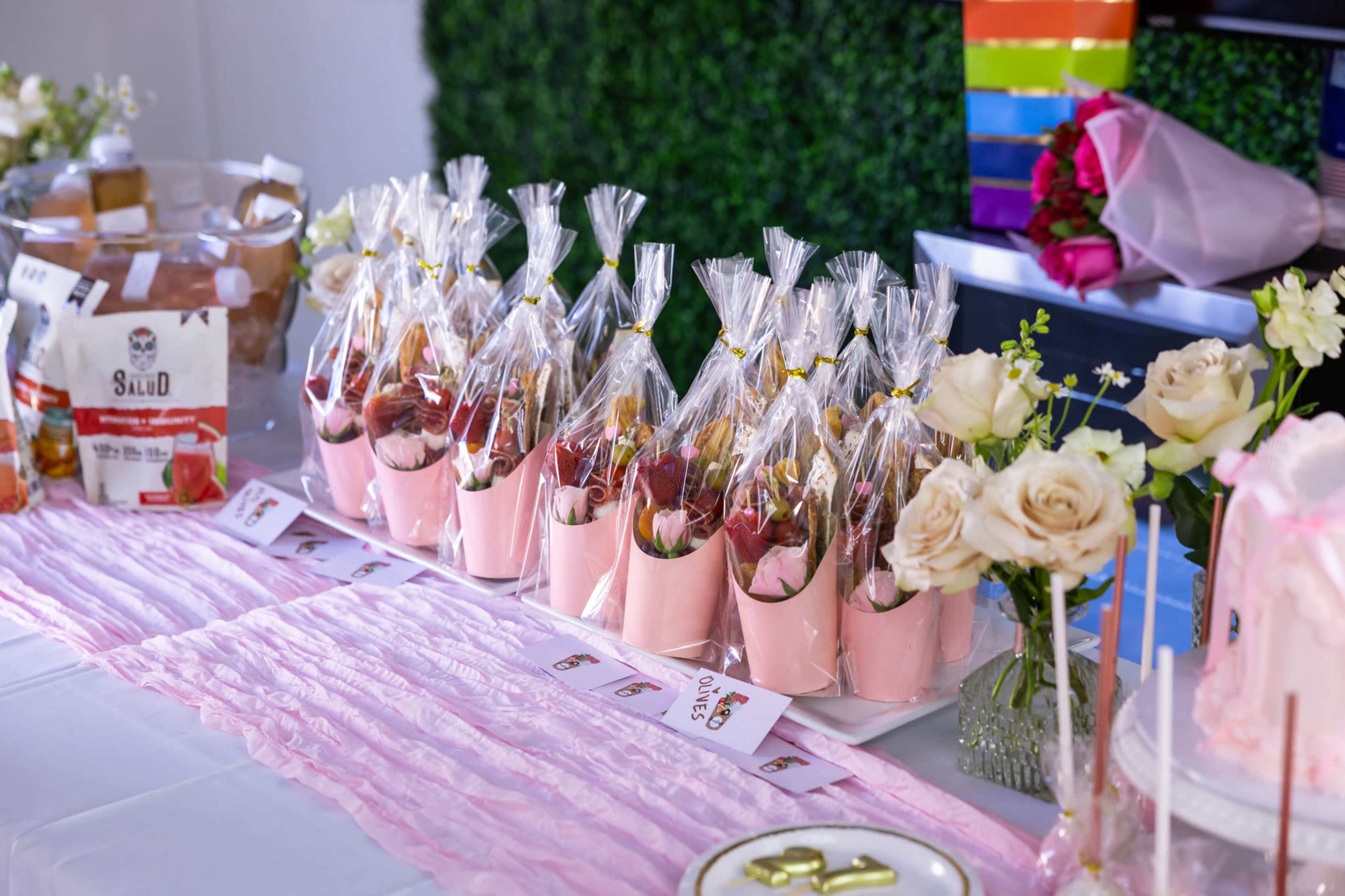 The image shows a table set with neatly packaged treats in pink cups, surrounded by flowers and decorative items.