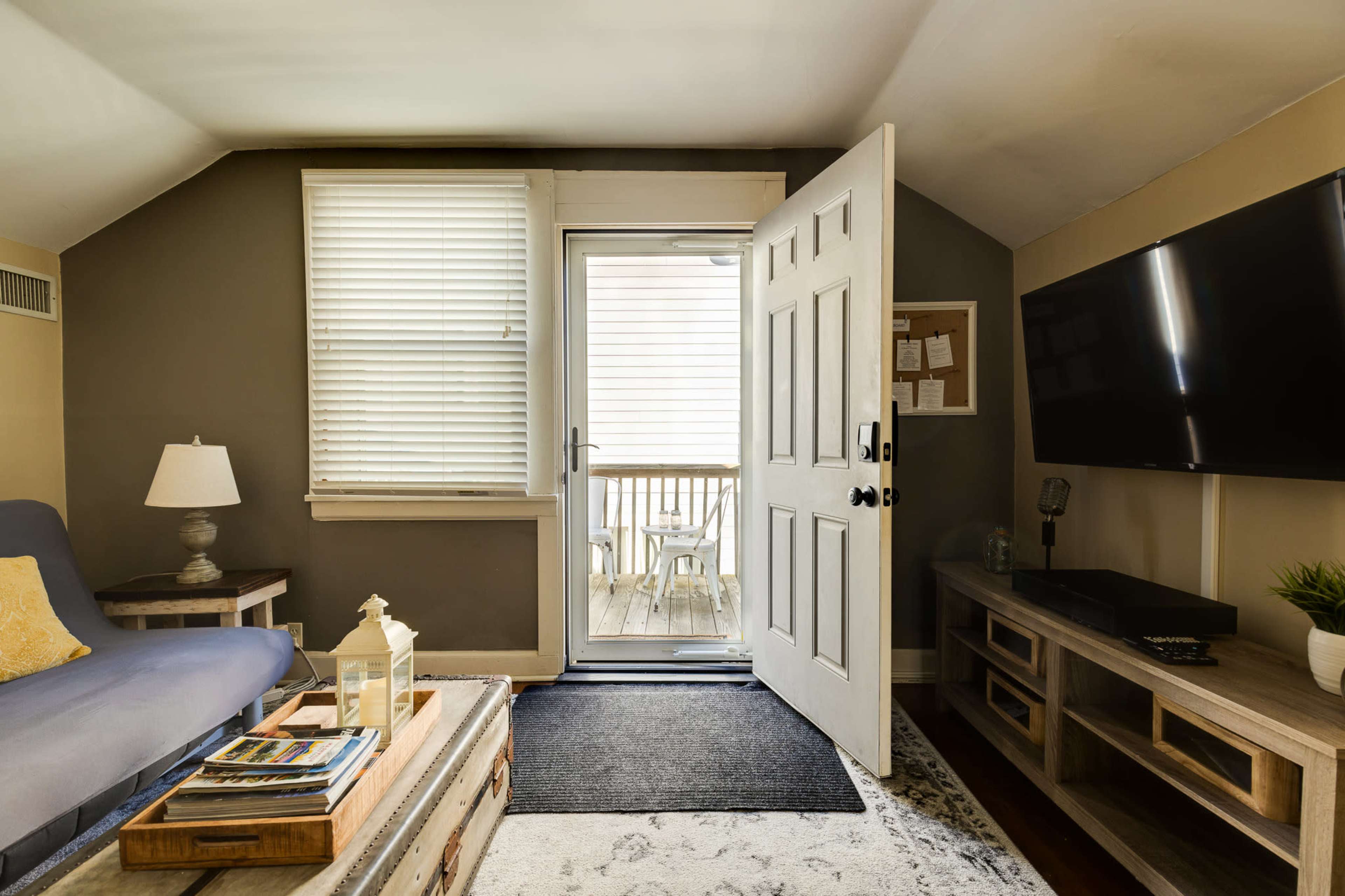 The image shows a cozy living room with an open door leading to a small deck area, featuring a blue sofa, a coffee table with magazines, and a TV mounted on the wall.