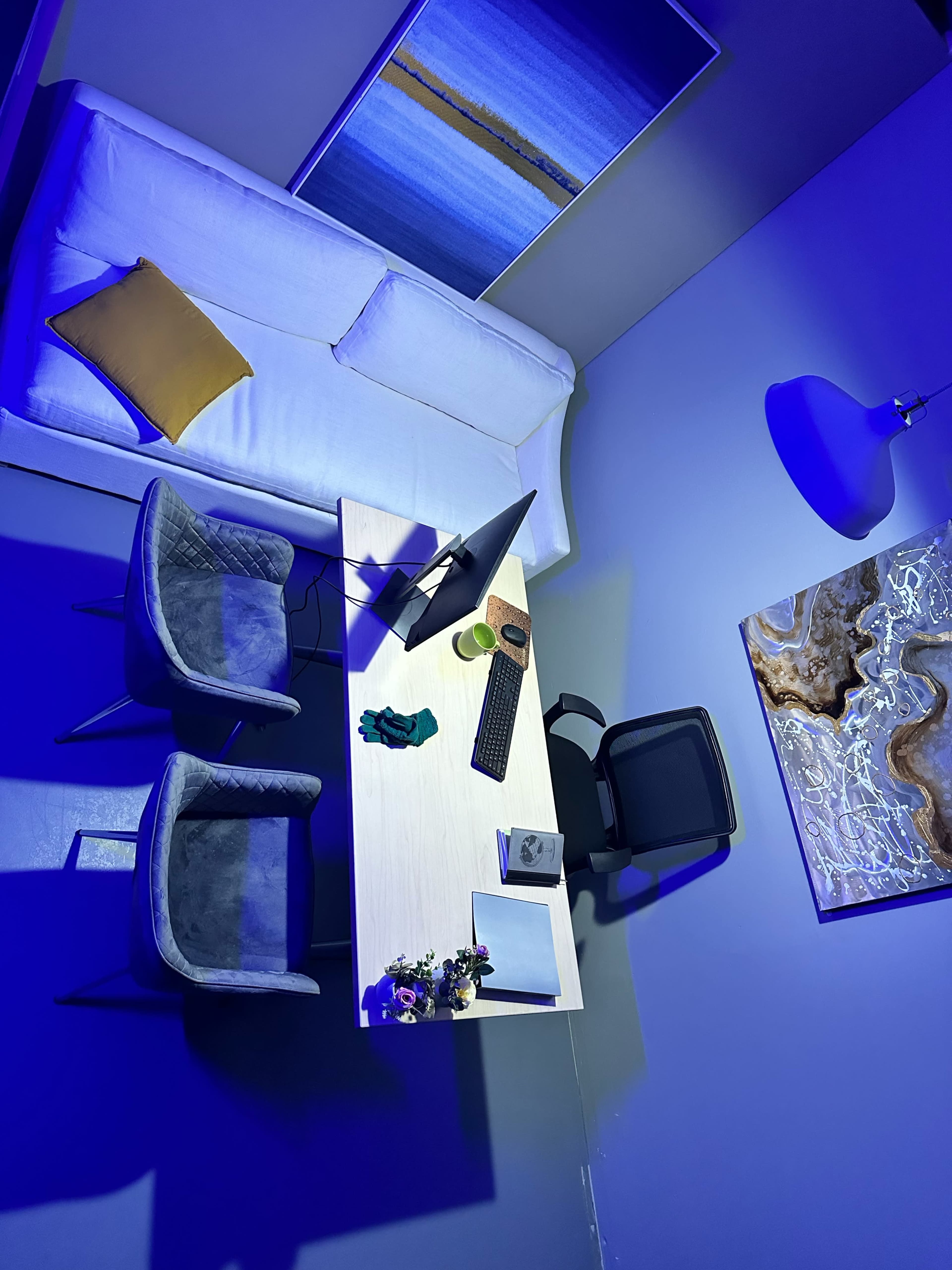 A modern workspace featuring a white sofa, a wooden desk with a computer, two gray chairs, and decorative elements against blue lighting.