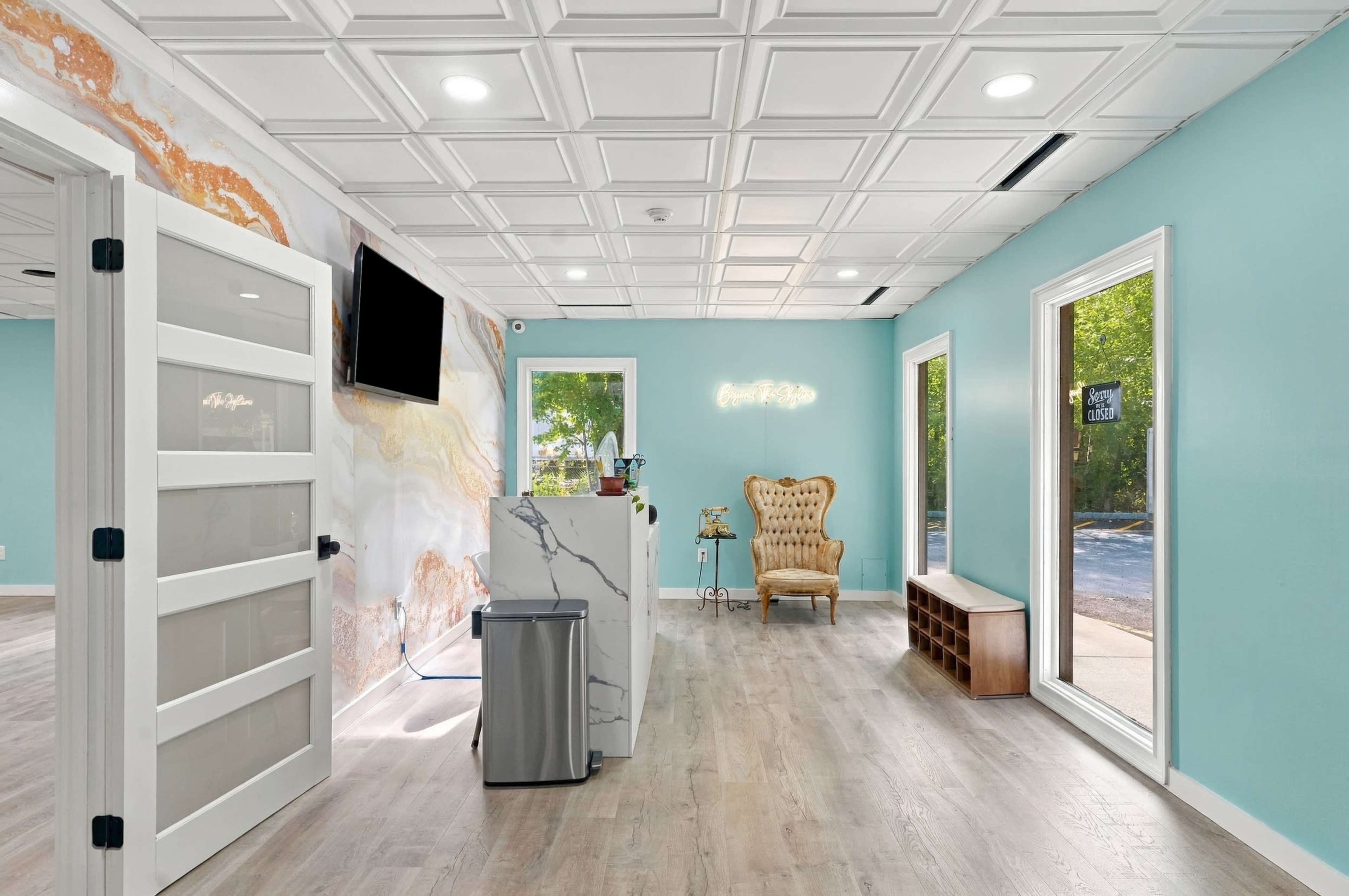 The image shows a bright, modern reception area with teal walls, a marble counter, a plush chair, and large windows.