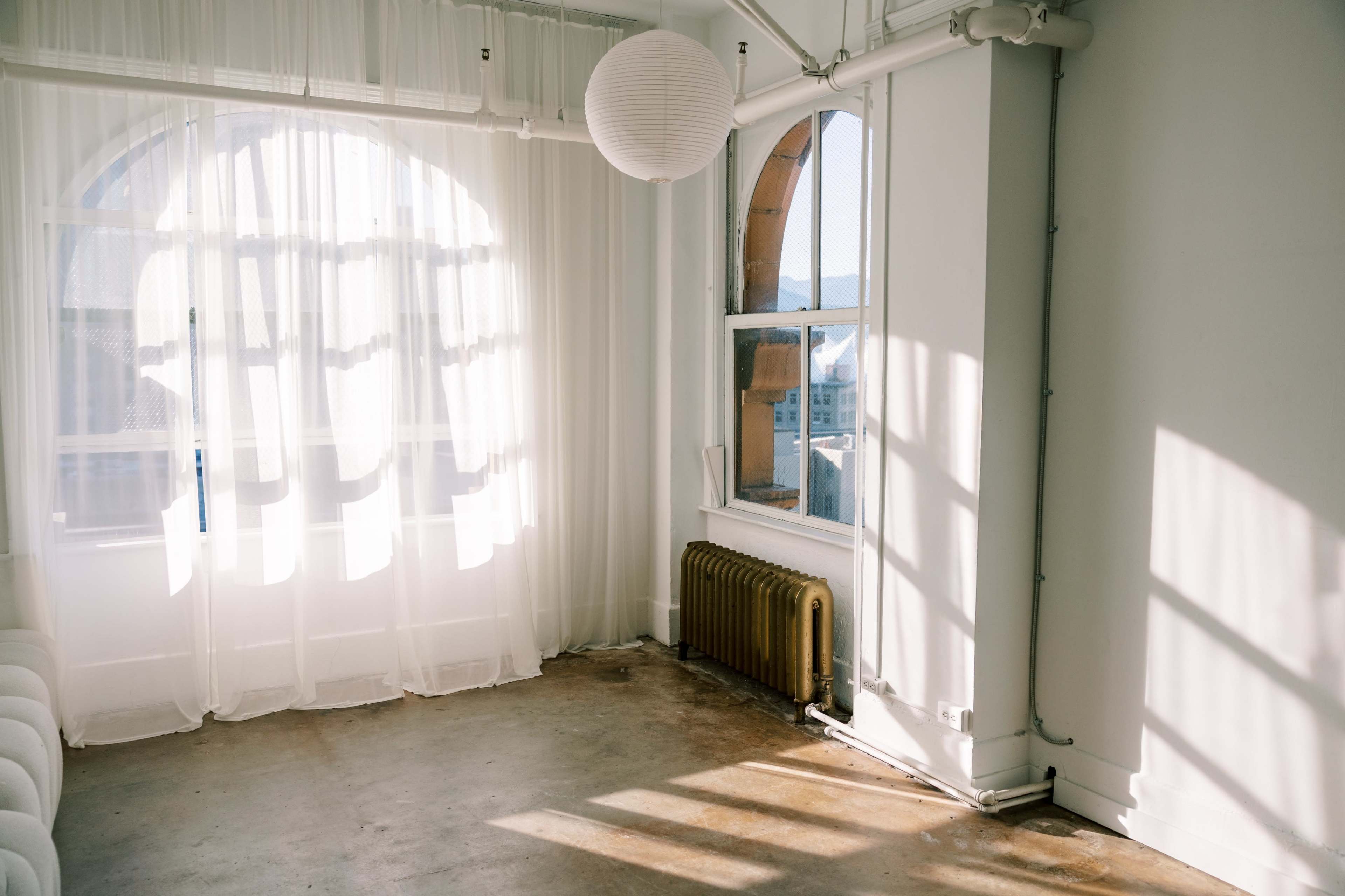 A bright, airy room features large windows with sheer curtains, casting shadows on the polished concrete floor.