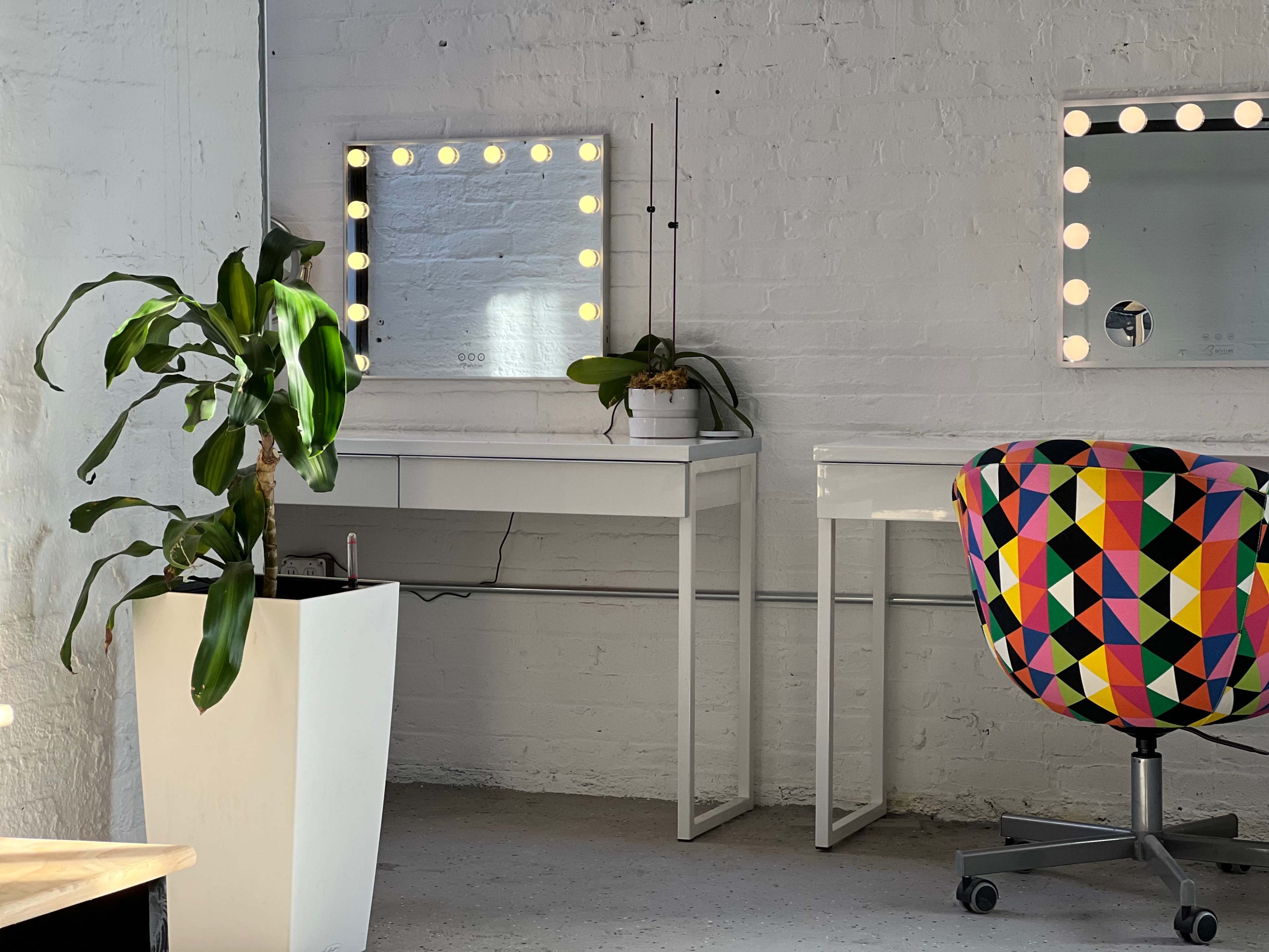A brightly lit room features a white vanity desk with a mirror surrounded by light bulbs, a colorful patterned chair, and a potted plant beside it.