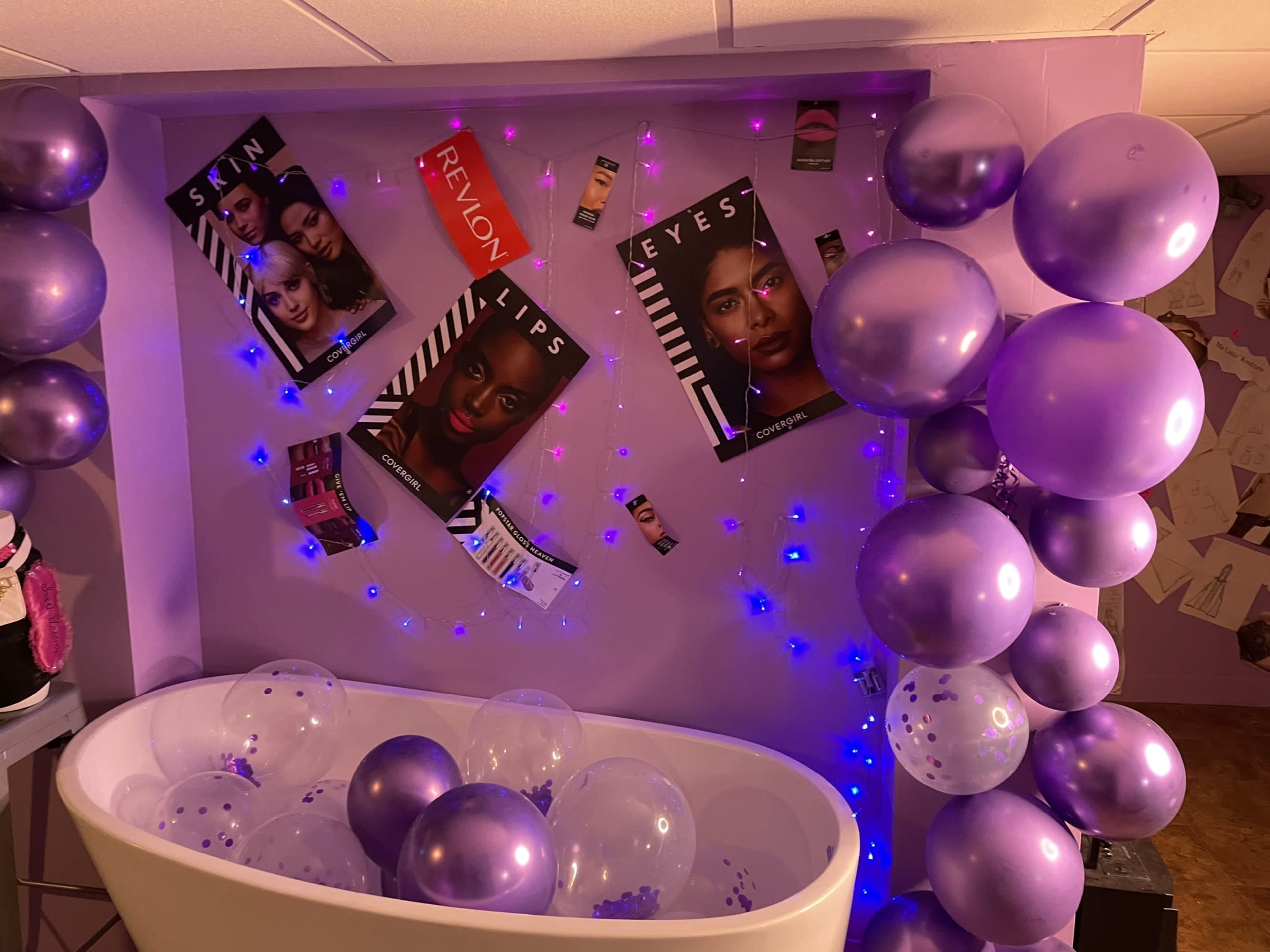The image features a decorated bathroom area with a bathtub filled with clear balloons, purple balloons, and magazine posters displayed on the walls.