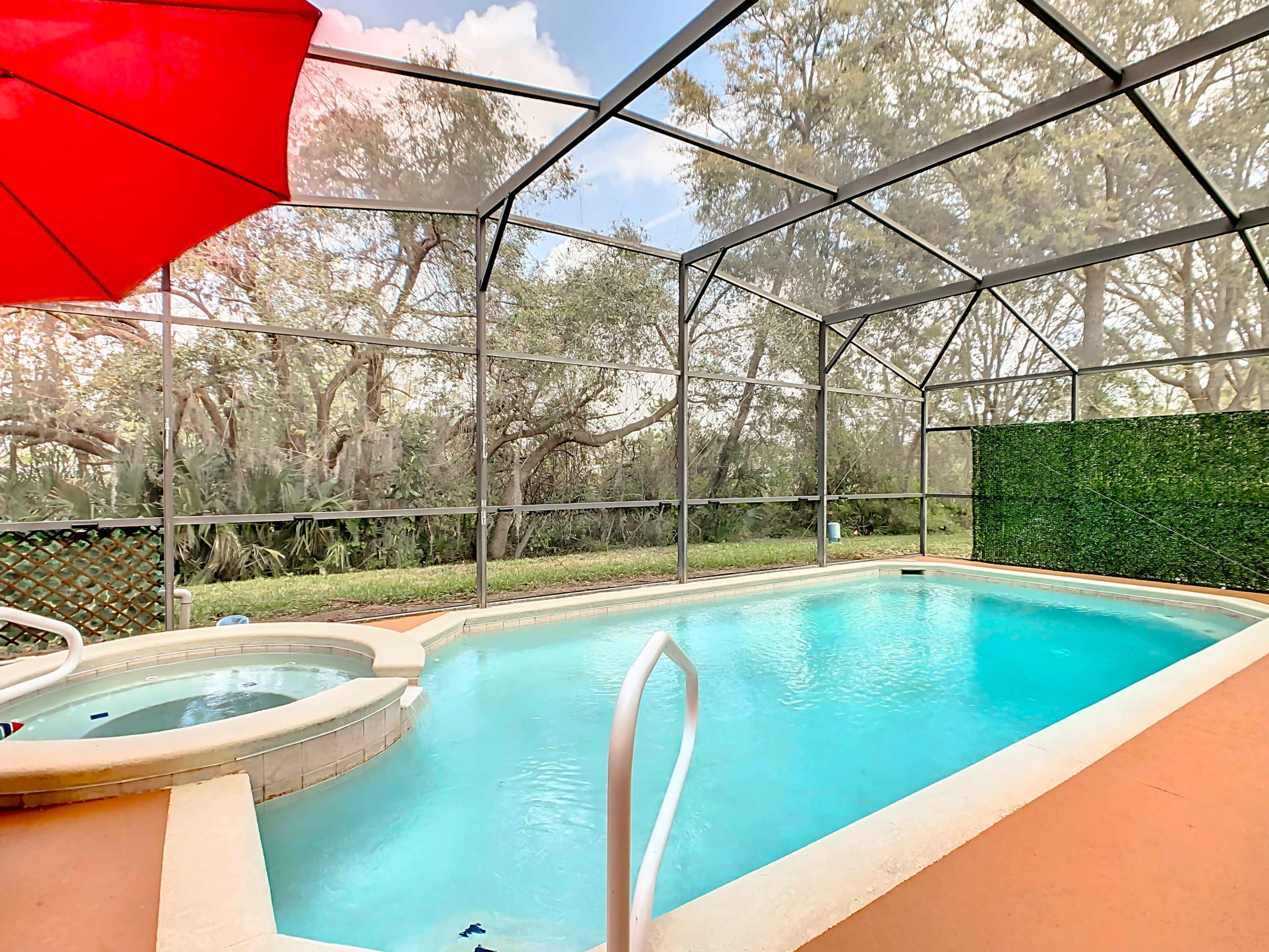 A swimming pool with an attached spa is enclosed in a screened patio surrounded by greenery and trees.