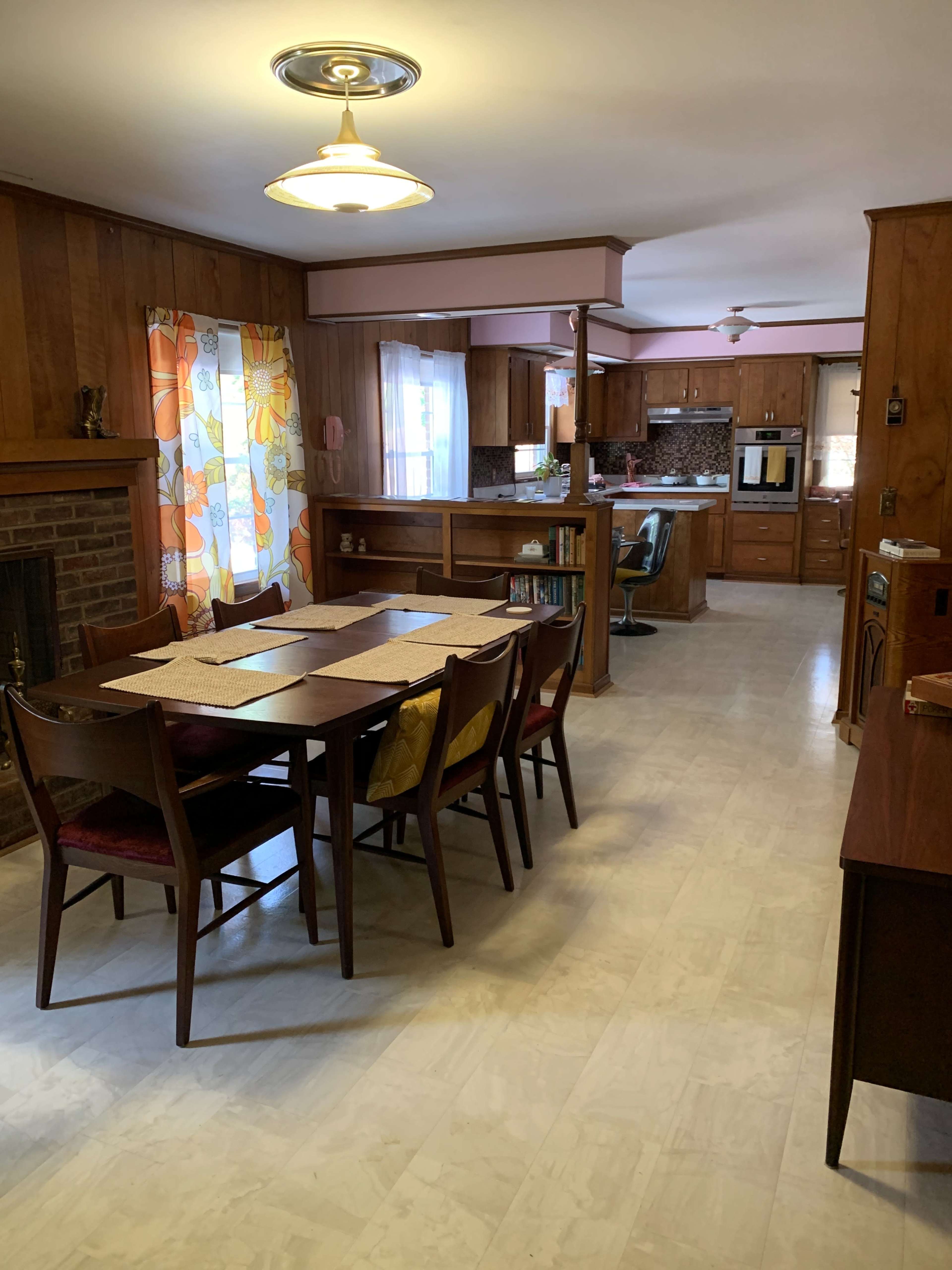 The image shows a dining area with a wooden table and chairs, leading into a kitchen with wooden cabinetry and floral curtains.