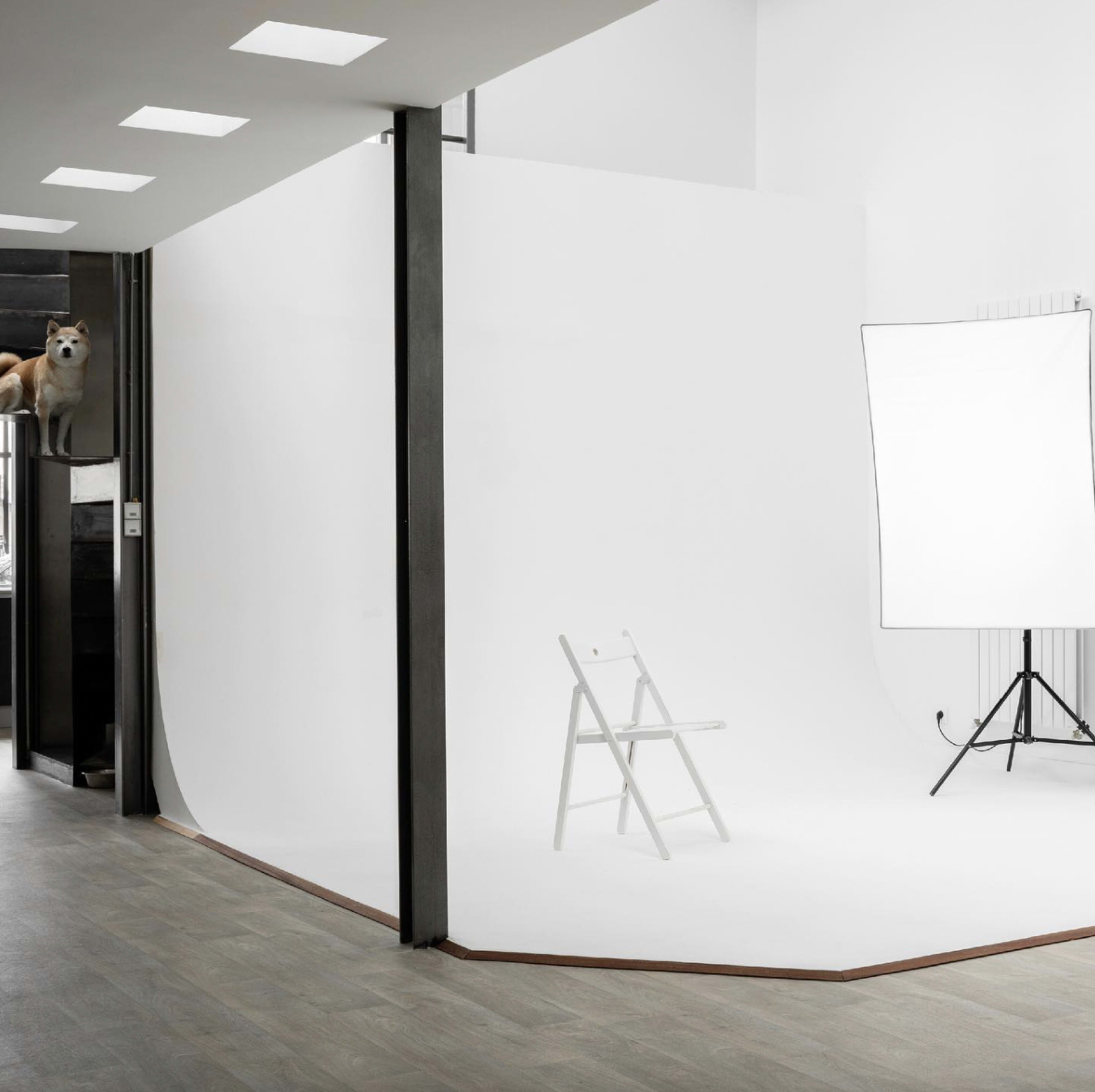 A minimalist studio space features a white backdrop with a single white chair and a spotlight setup, alongside an open doorway.